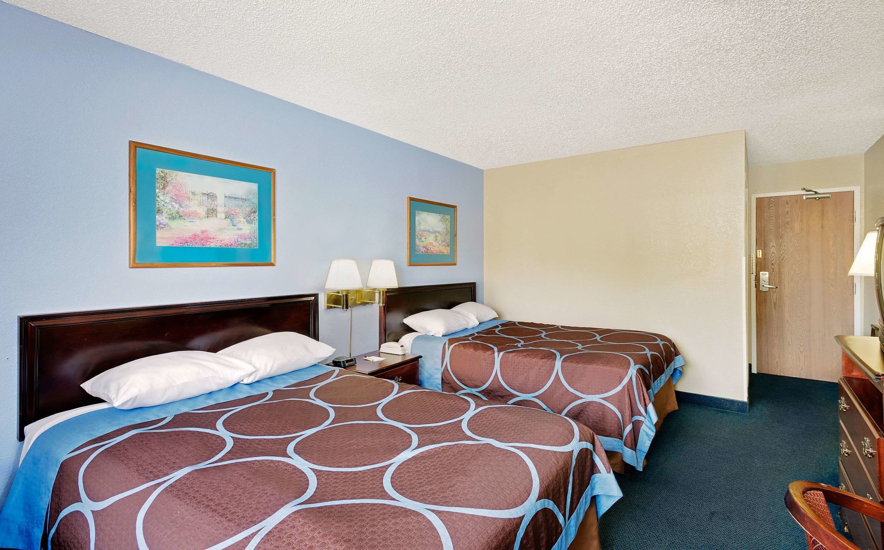 A hotel room with two beds, each with a brown and blue patterned bedspread, a wooden headboard, a nightstand with a lamp, and two framed artworks on the wall.
