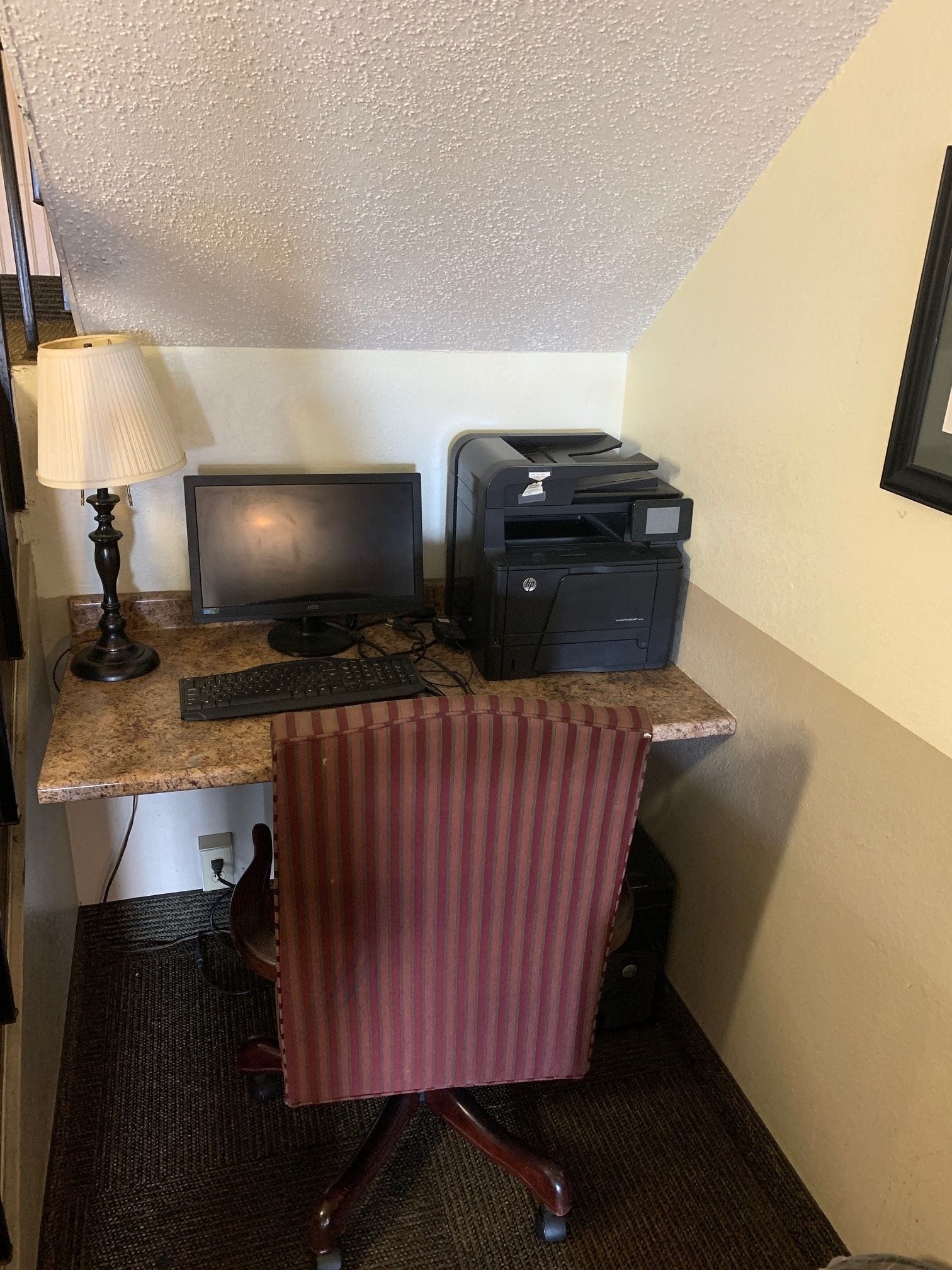 Une chambre d’hôtel compacte équipée d’un bureau, d’un écran d’ordinateur, d’une imprimante et d’une chaise à rayures.