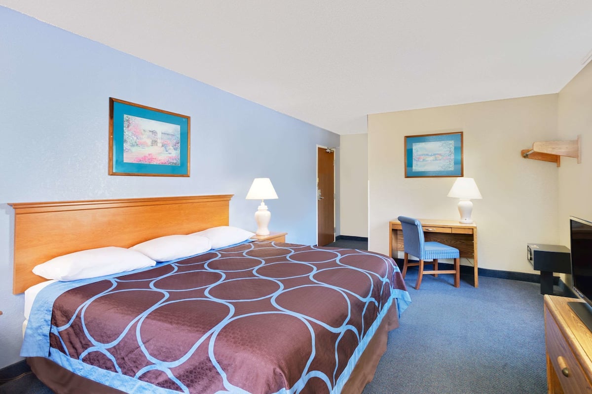 Ein Hotelzimmer mit einem großen Bett, einem Schreibtisch mit Stuhl, einem Fernseher und zwei gerahmten Bildern an den Wänden.