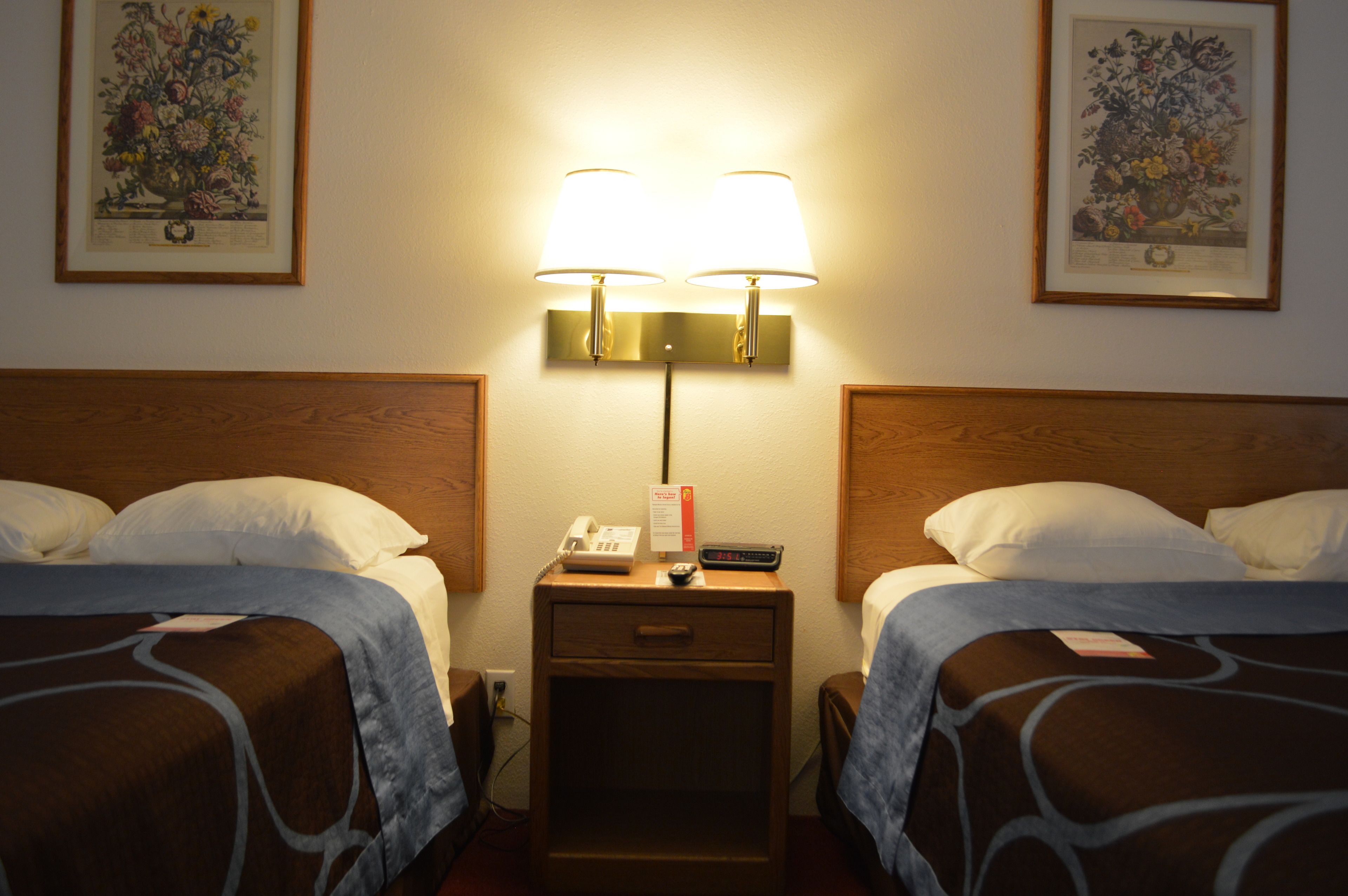A hotel room with two beds, a nightstand with a lamp, a phone, and a remote control.