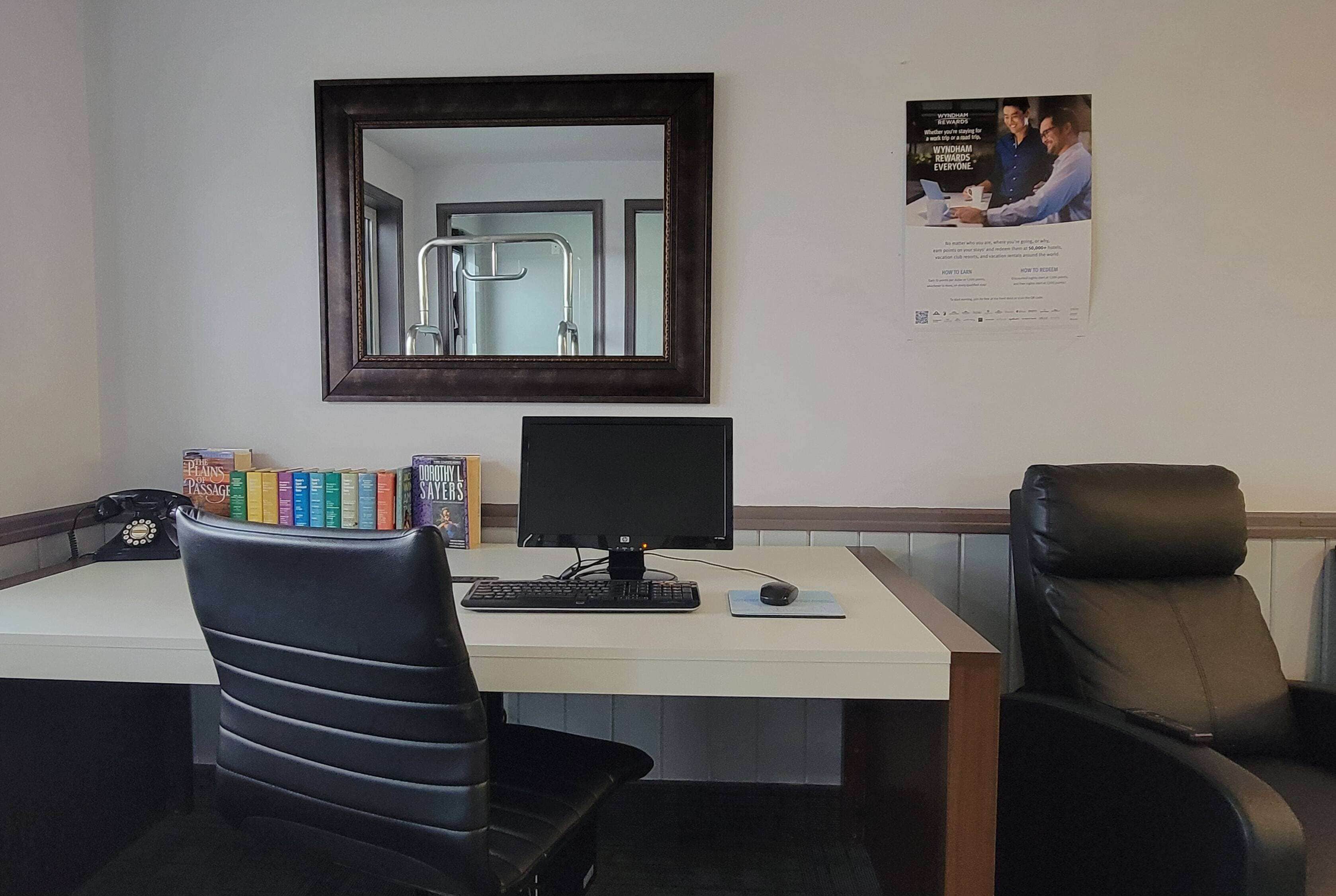 A desk with a computer, a chair, and a mirror. There are books on the desk and a poster on the wall.