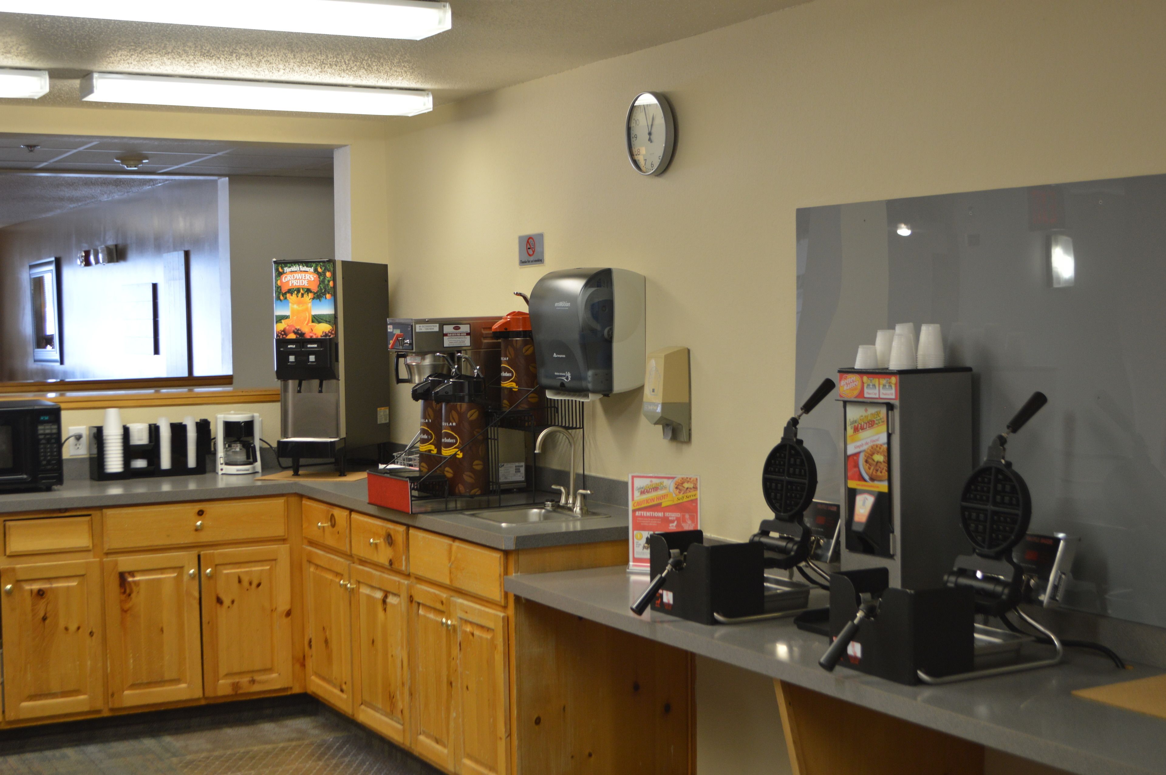A kitchen area with wooden cabinets, a coffee machine, waffle makers, and a clock on the wall.