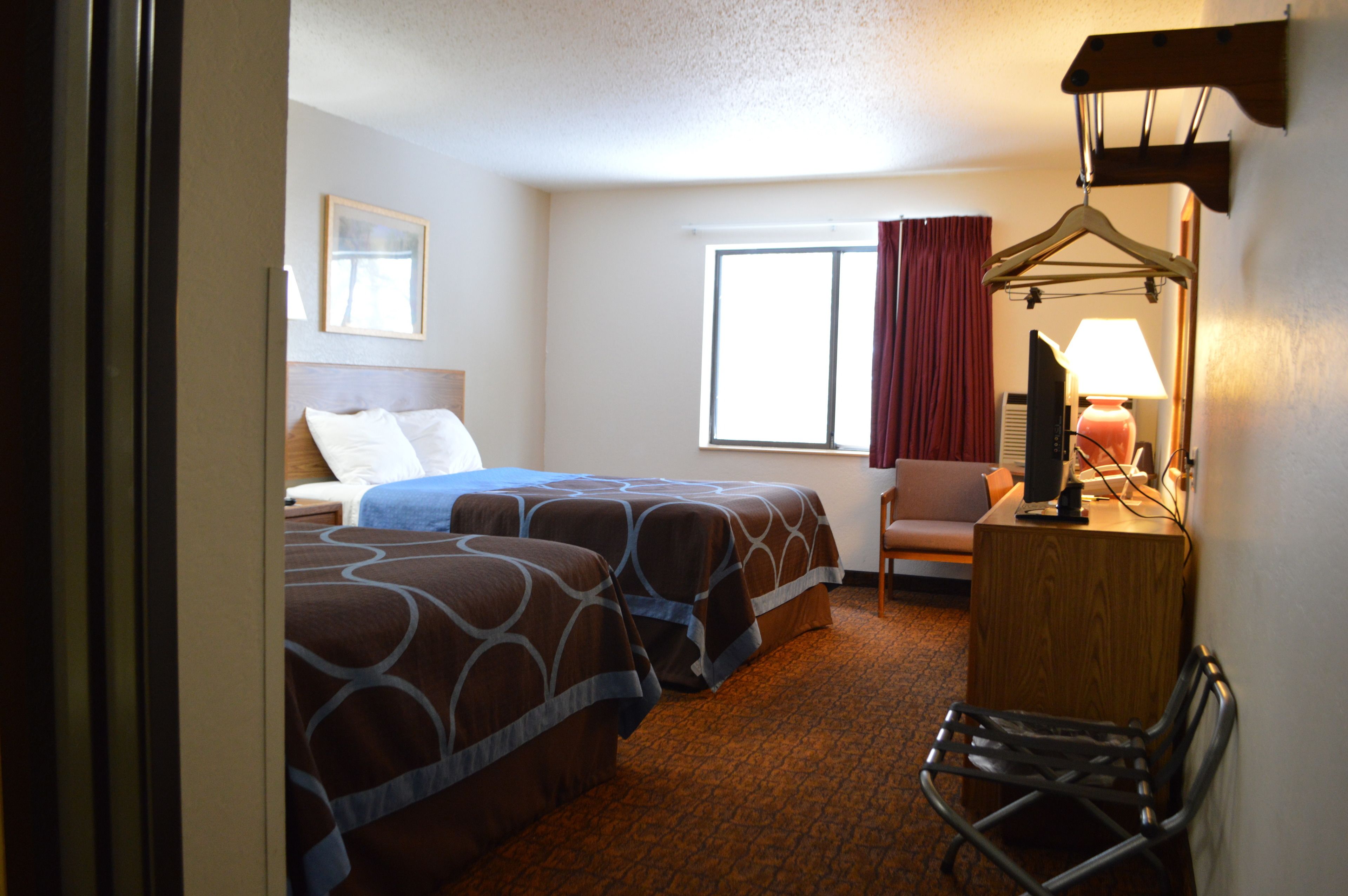 A hotel room with two beds, a desk with a lamp, a chair, a window with curtains, and a framed picture on the wall.