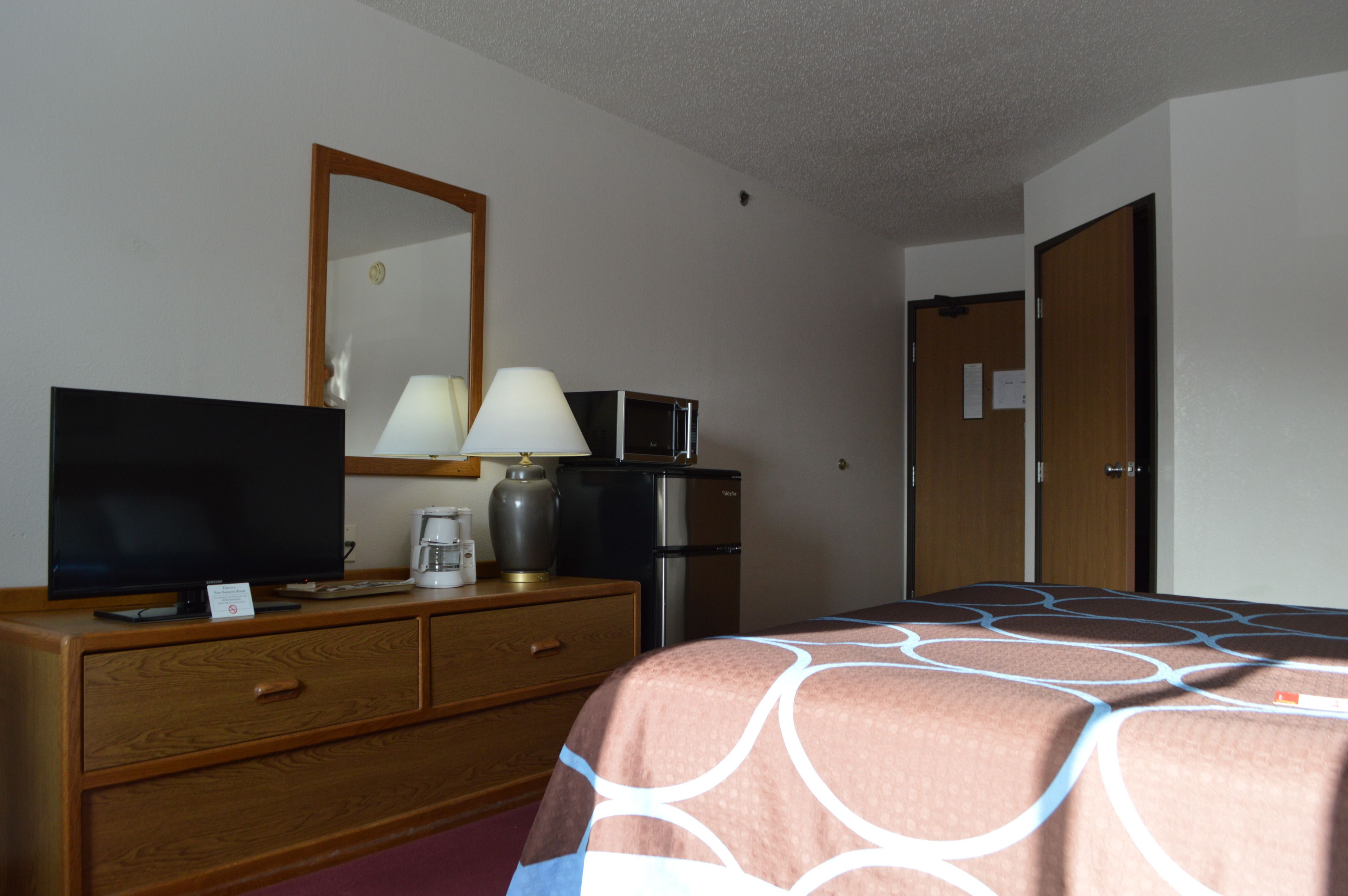 A hotel room with a bed, a TV, a lamp, a coffee maker, a refrigerator, and a wooden dresser.