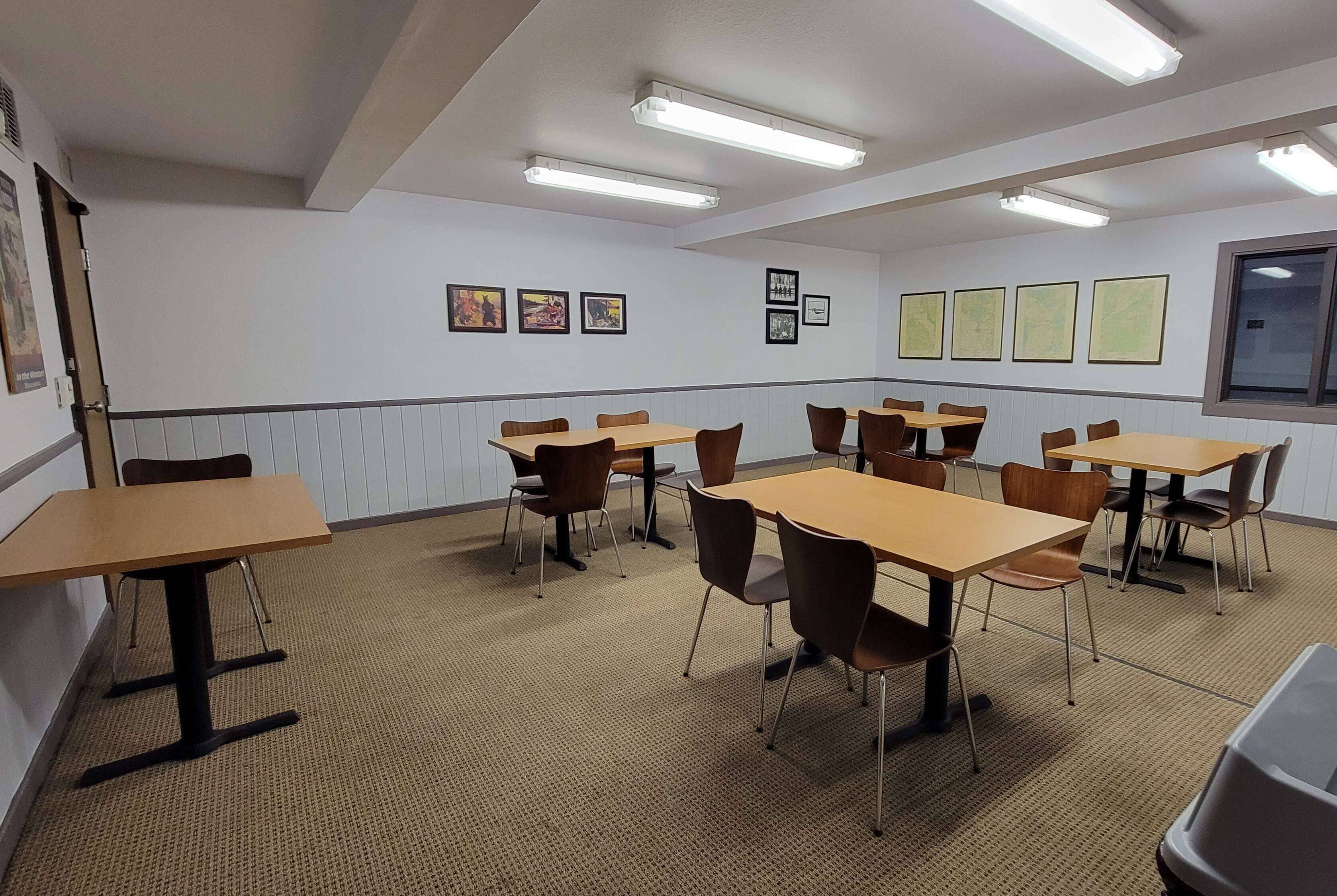 A conference room with tables and chairs, fluorescent ceiling lights, and framed pictures on the walls.