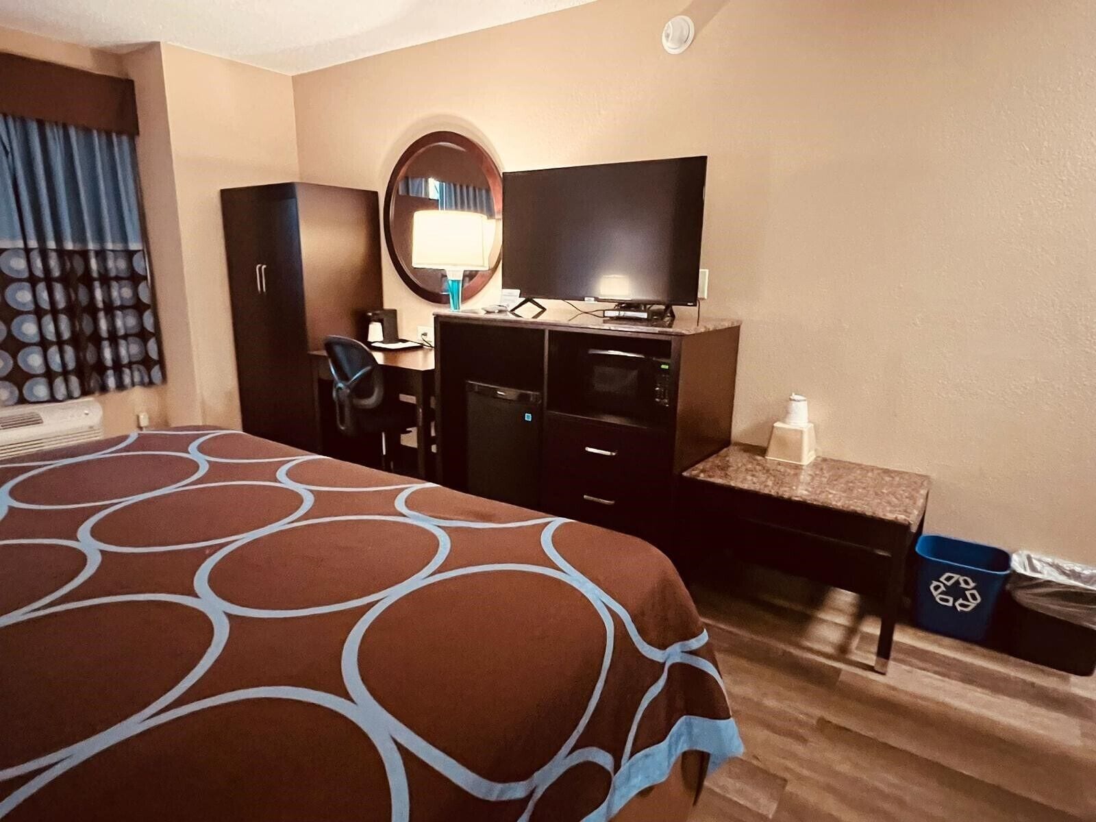 A hotel room with a bed, a desk with a computer, a TV, a microwave, a small trash bin, and a recycling bin.