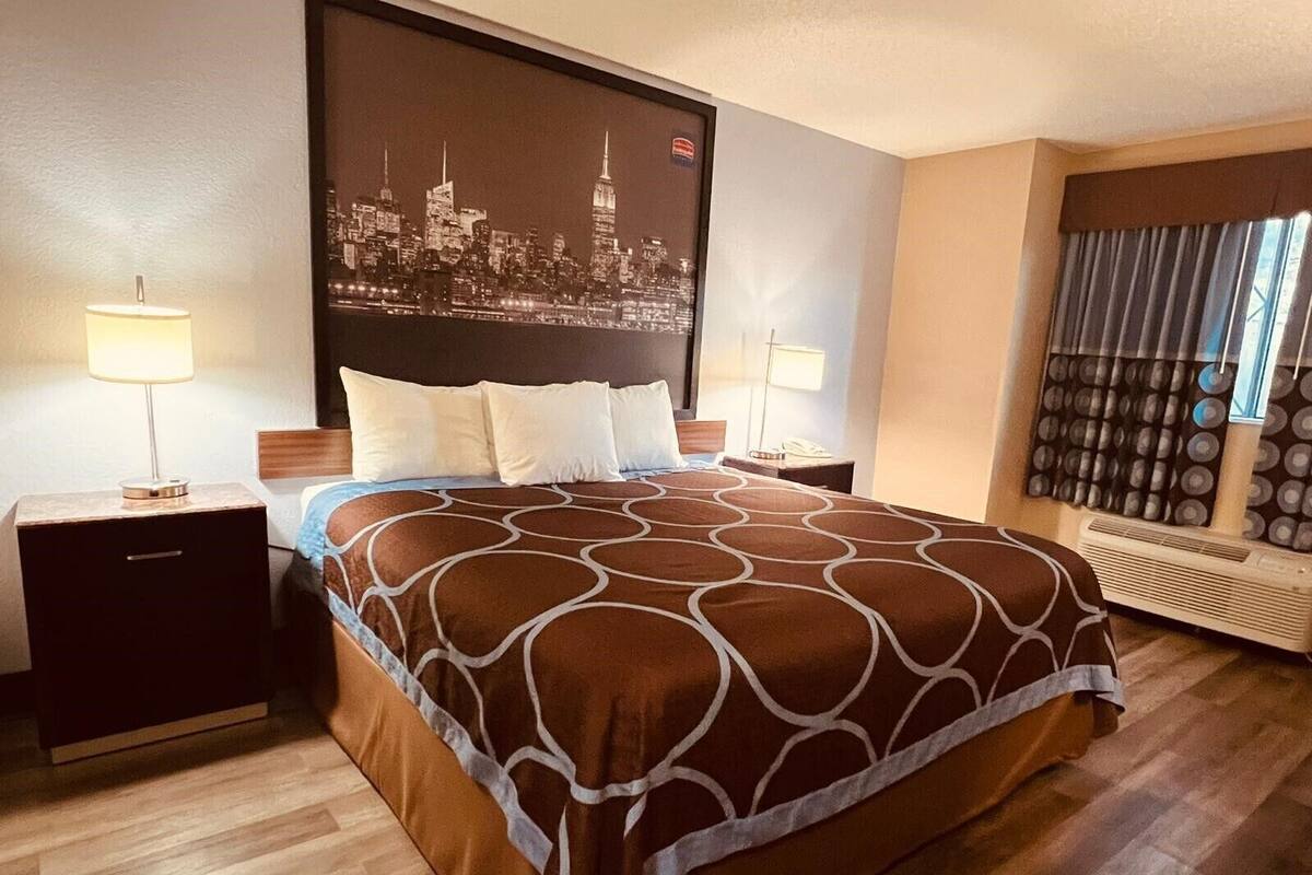 A hotel room with a large bed, two bedside tables with lamps, a cityscape wall mural, a window with curtains, and a wall-mounted air conditioning unit.