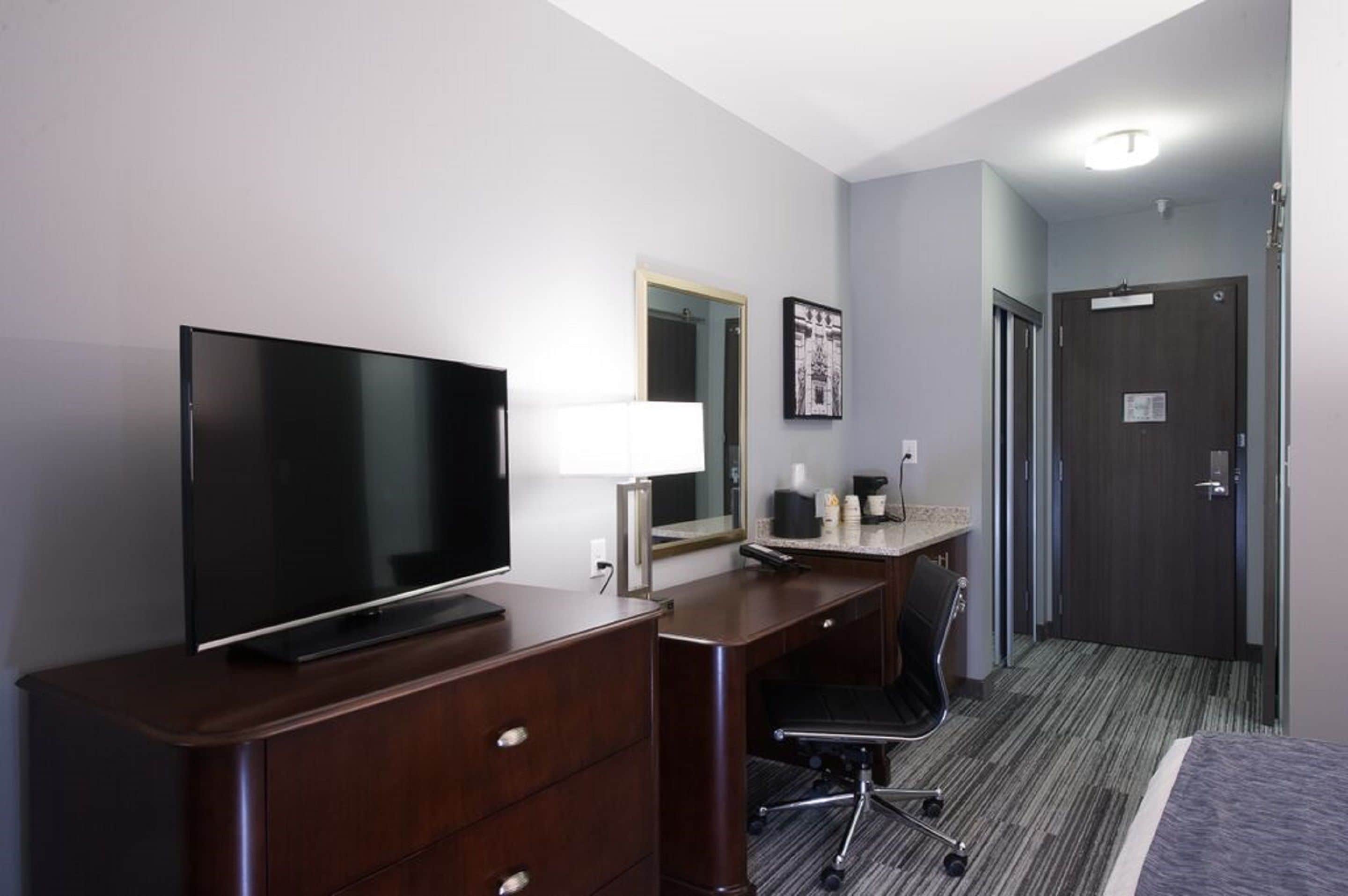 A hotel room with a flat-screen TV, a desk with a lamp, a chair, and a bed.