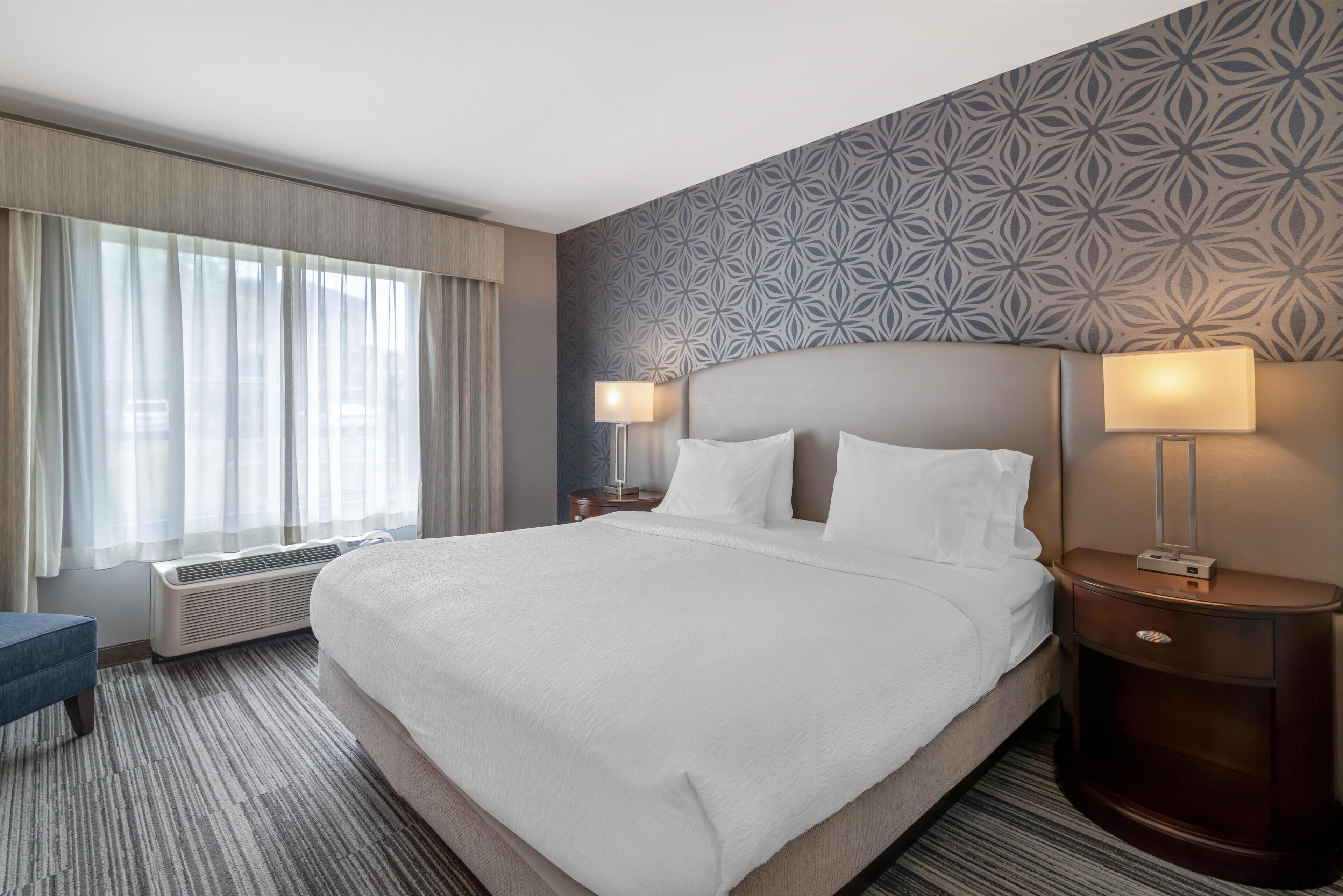A hotel room with a large bed, two bedside lamps, a nightstand, a window with curtains, and a patterned wall.