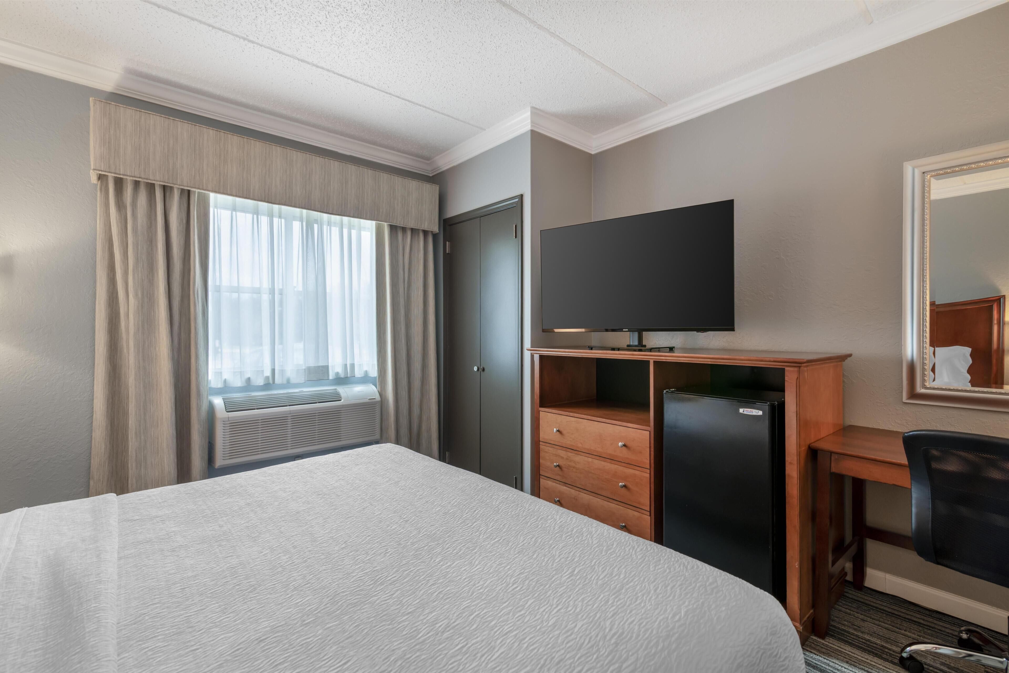 A hotel room with a bed, a TV on a wooden stand, a desk with a chair, and a large window with curtains.