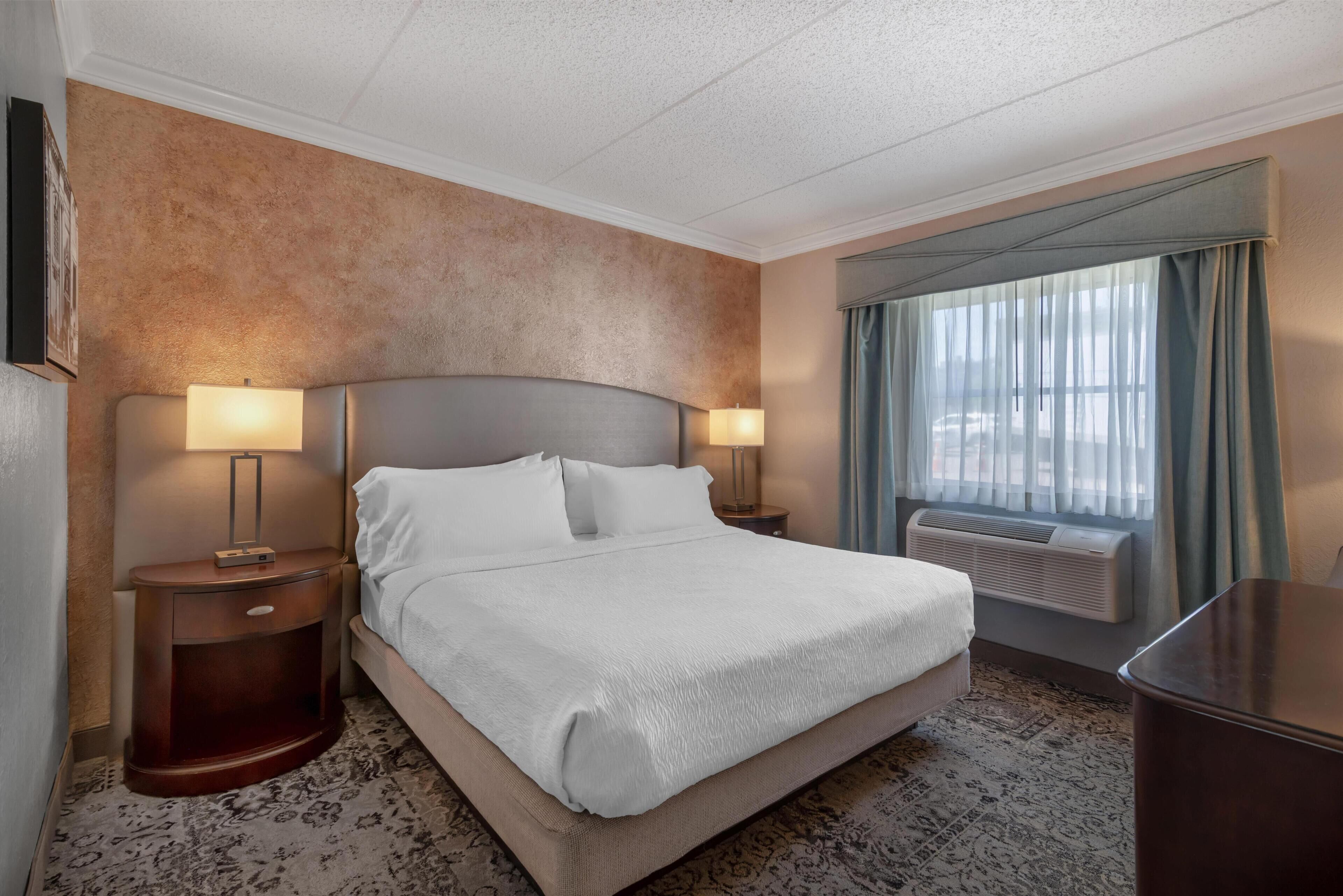 A hotel room with a large bed, two bedside lamps, a window with curtains, and a wall-mounted air conditioner.