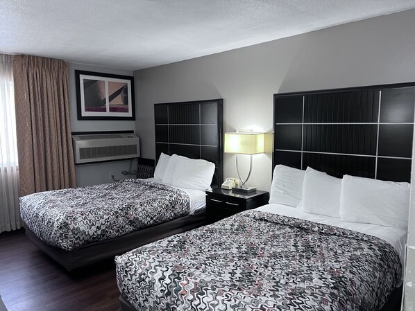 A hotel room with two beds, a nightstand with a lamp, a framed picture on the wall, and an air conditioning unit.