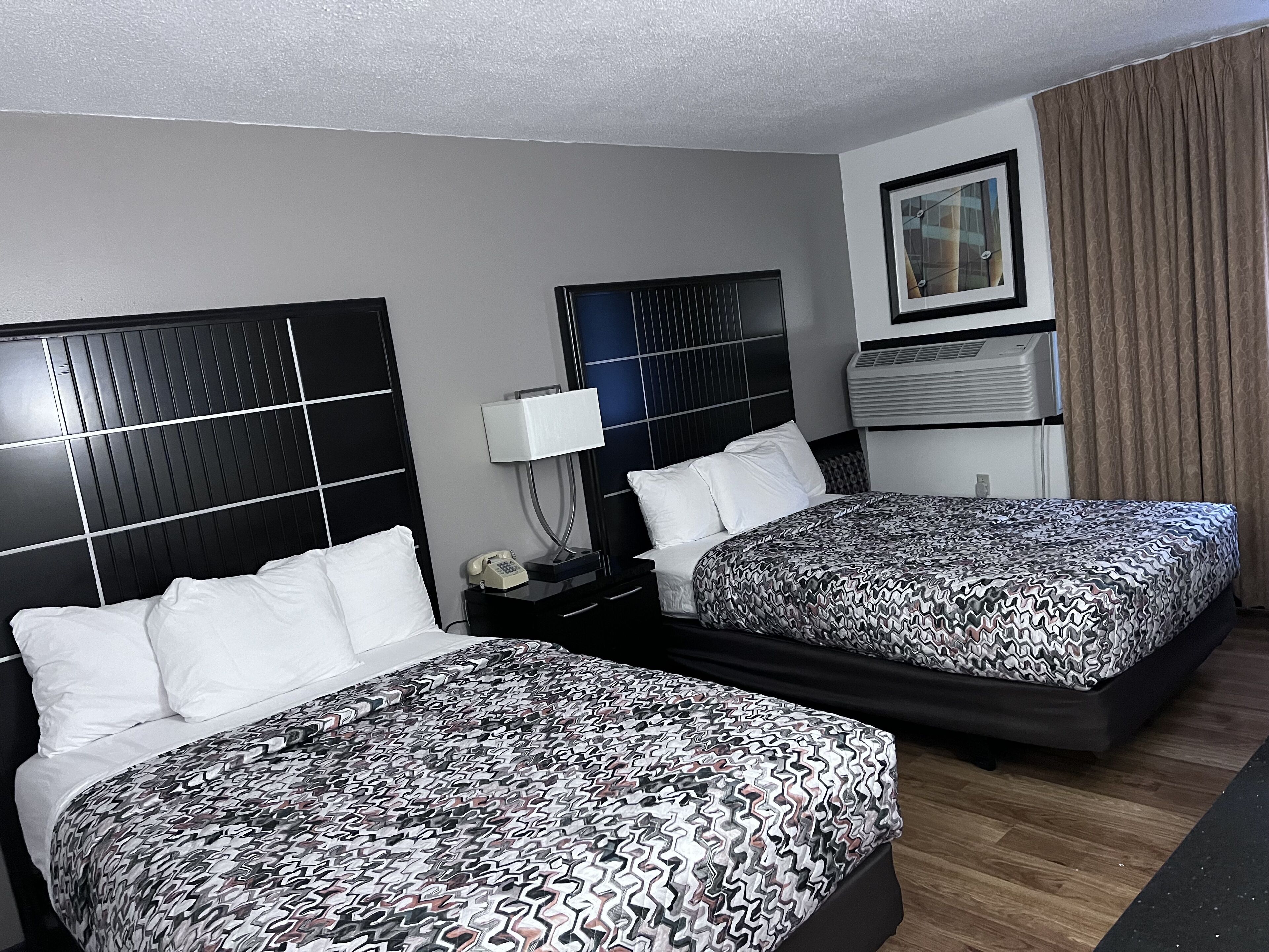 A hotel room with two beds, a bedside table with a lamp, a phone, and a wall-mounted air conditioning unit.
