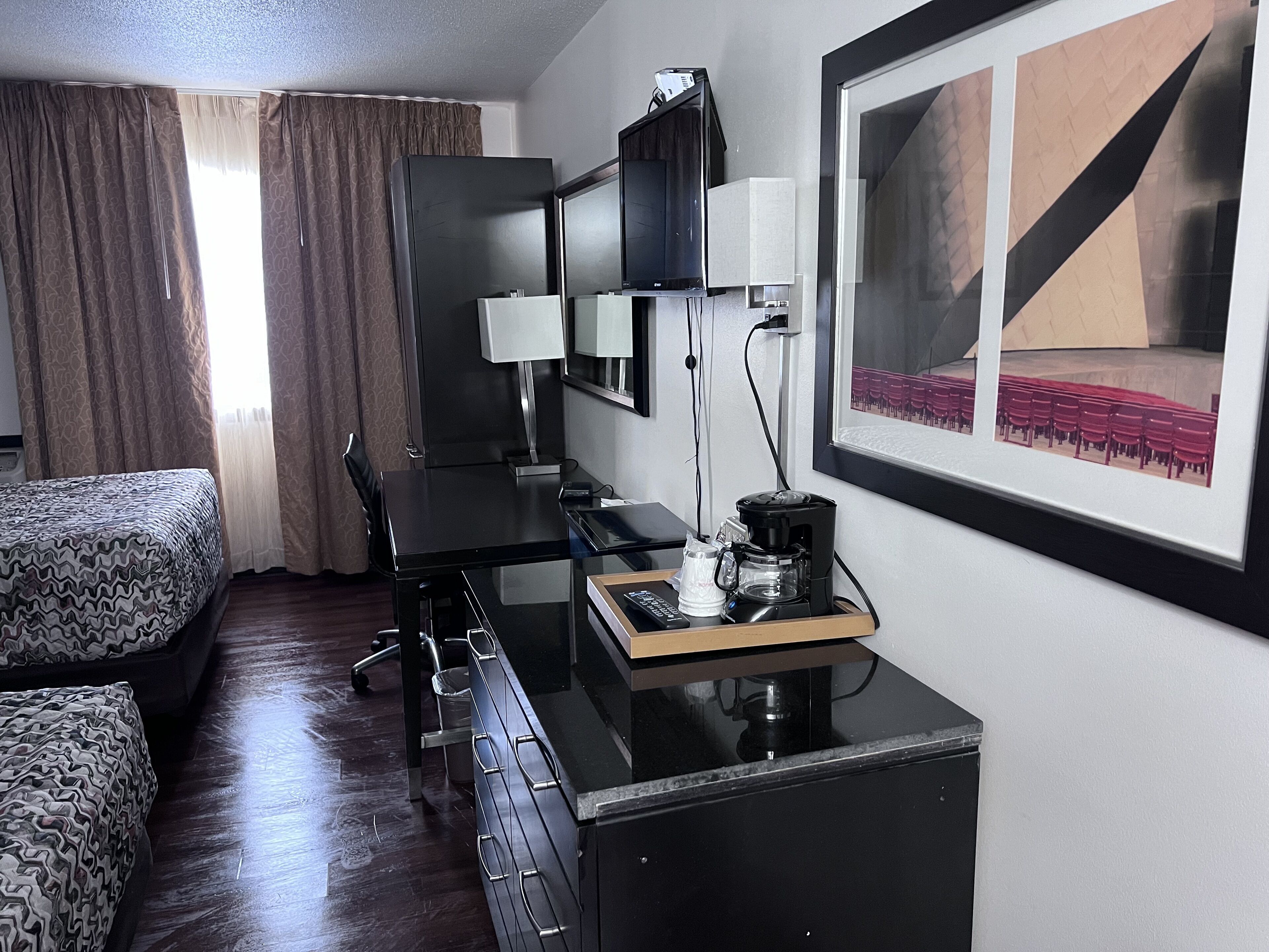A hotel room with a bed, a desk with a computer, a coffee maker, and a framed picture on the wall.
