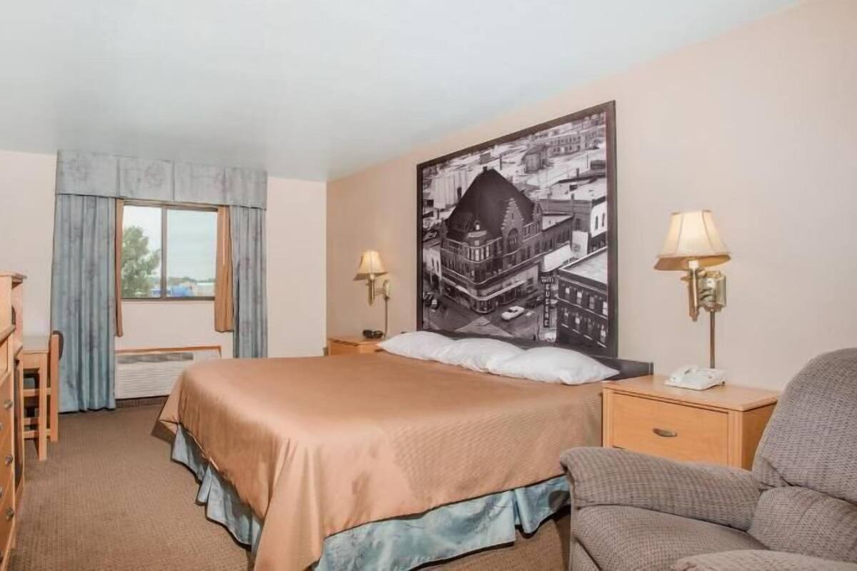 Une chambre d’hôtel avec un lit, un bureau, une chaise, une télévision, une fenêtre avec des rideaux et un grand tableau représentant un paysage urbain accroché au mur.