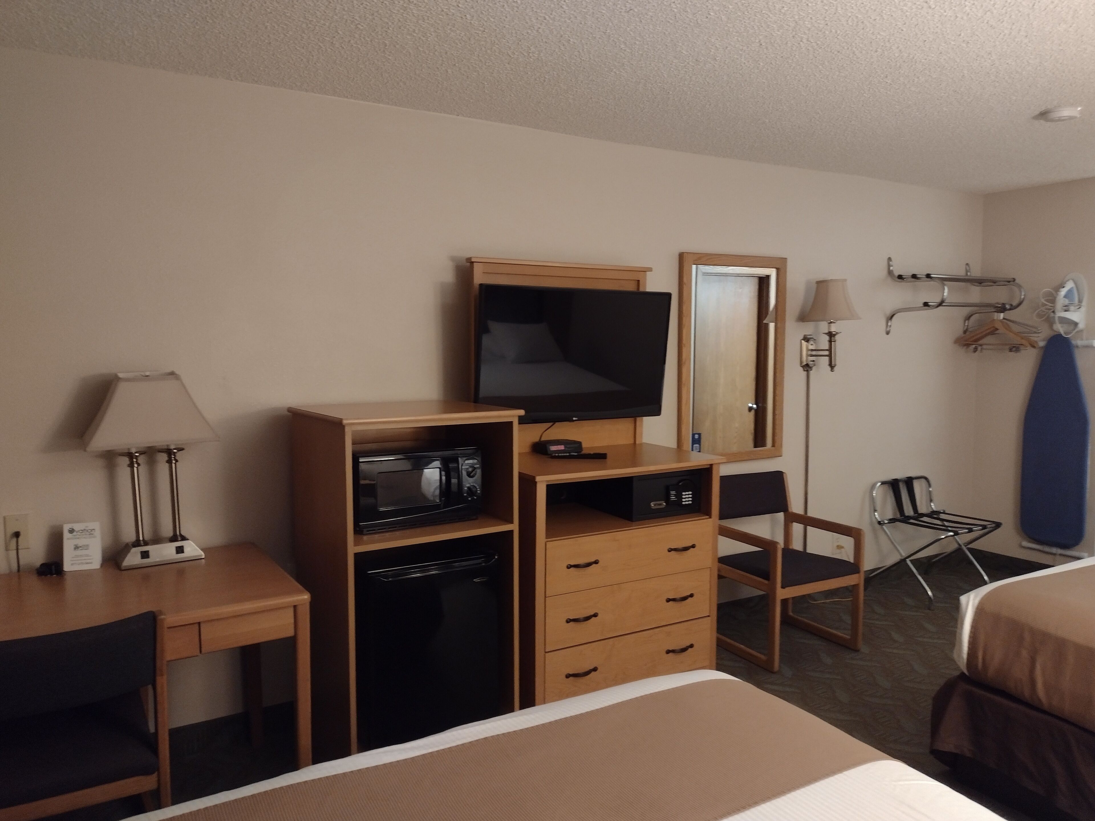 A hotel room with a bed, a desk with a lamp, a microwave, a television, a dresser, a chair, and a mirror.