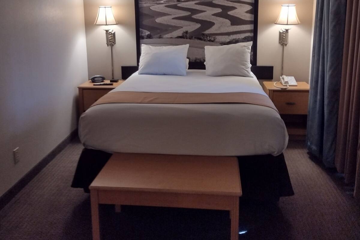 Une chambre d’hôtel avec un grand lit, deux lampes de chevet, une table de nuit, un banc et un grand tableau représentant un paysage accroché au mur.