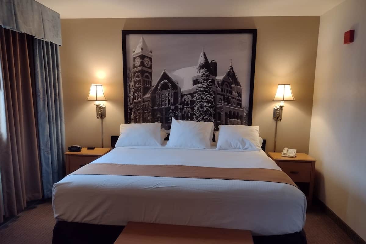 Une chambre d’hôtel avec un grand lit, deux lampes de chevet, un tableau encadré représentant un bâtiment historique et un banc en bois.