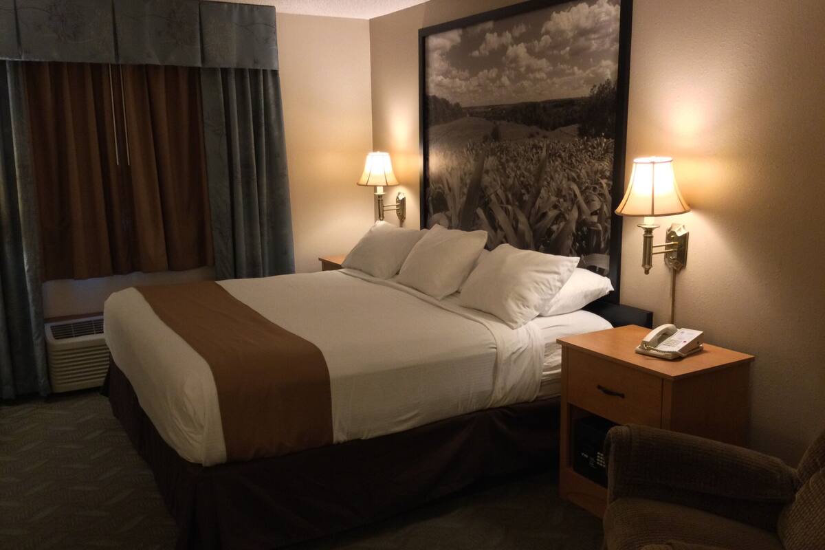Une chambre d’hôtel avec un grand lit, deux lampes de chevet, un téléphone, une chaise et un grand tableau représentant un paysage accroché au mur.