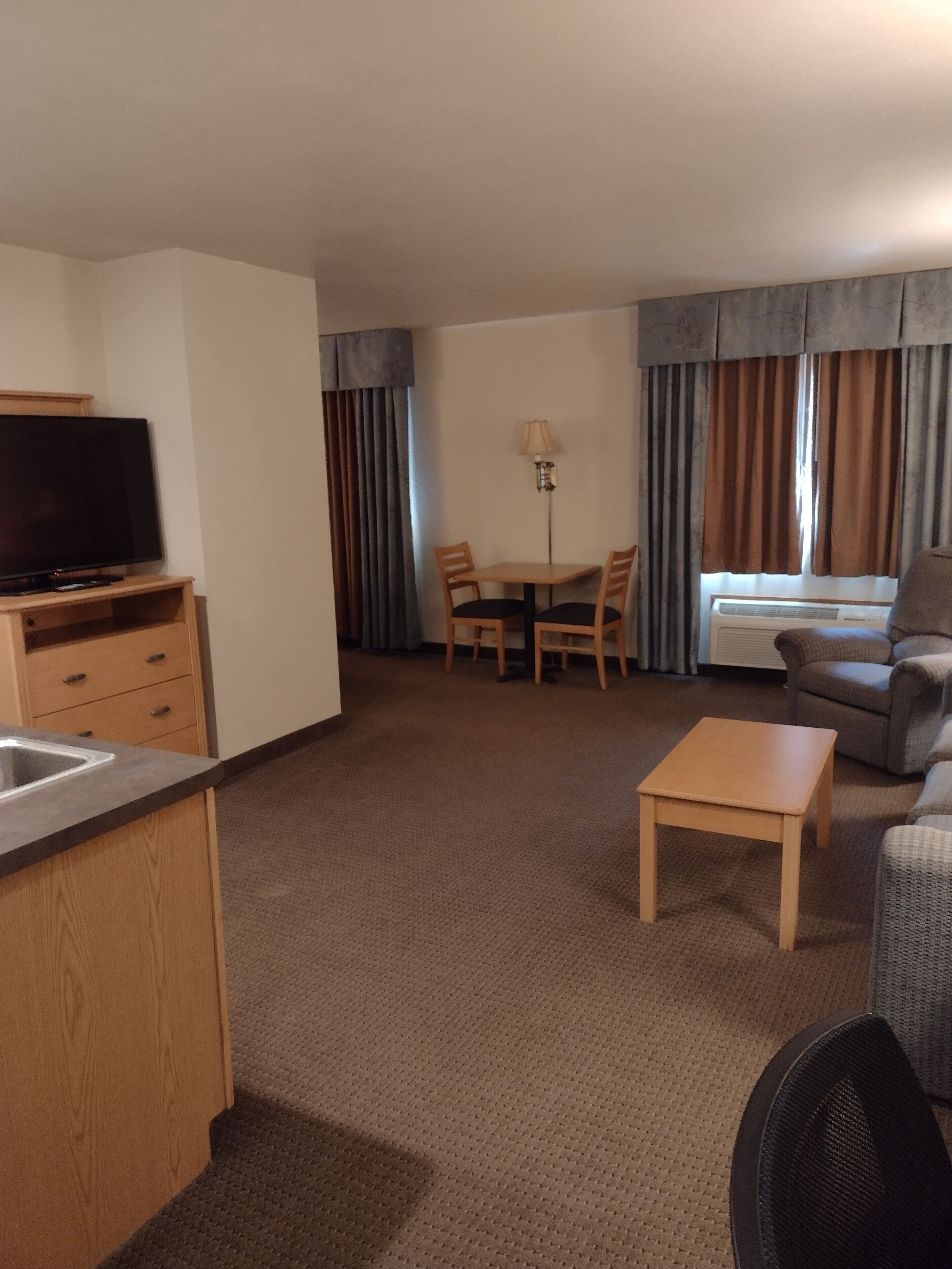 Ett kompakt hotellrum med ett litet kök, en platt-TV, en matplats med bord och stolar, en soffa och ett fönster med gardiner.