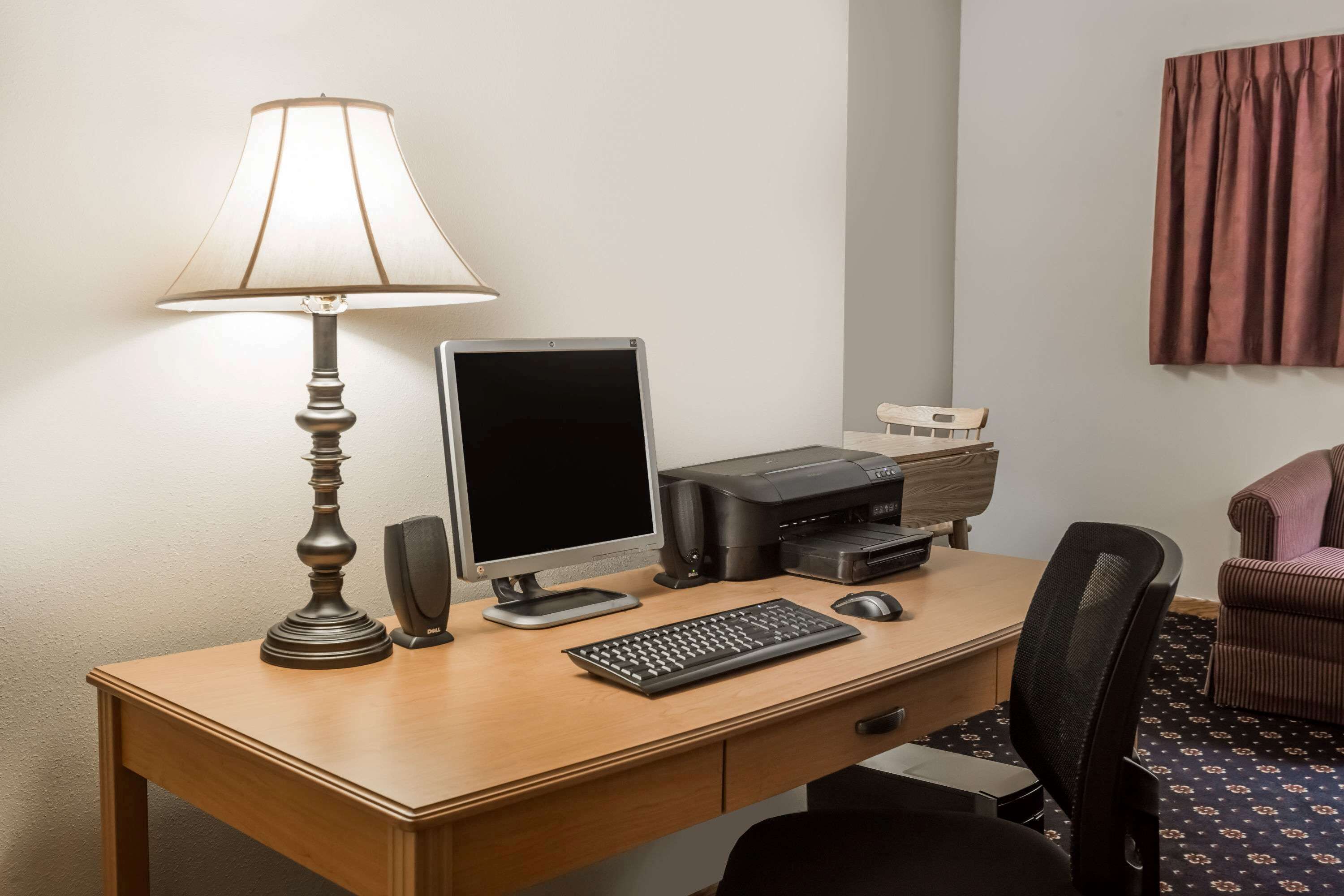 A hotel room with a wooden desk, a desktop computer, a lamp, a printer, and a chair.