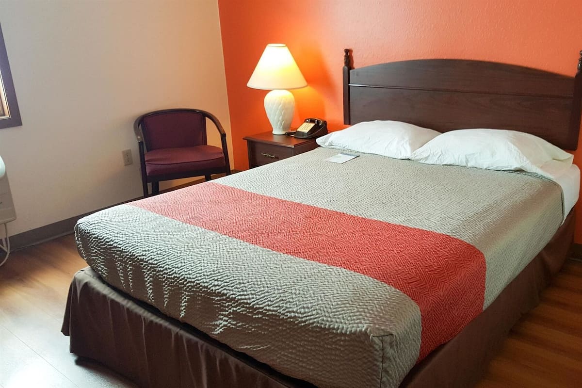 A neatly made bed with a wooden headboard, a bedside lamp, and a chair in a room with orange walls.