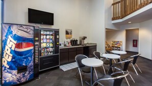 A modern hotel lobby with a vending machine, a flat-screen TV, a kitchenette, and a seating area with round tables and chairs.