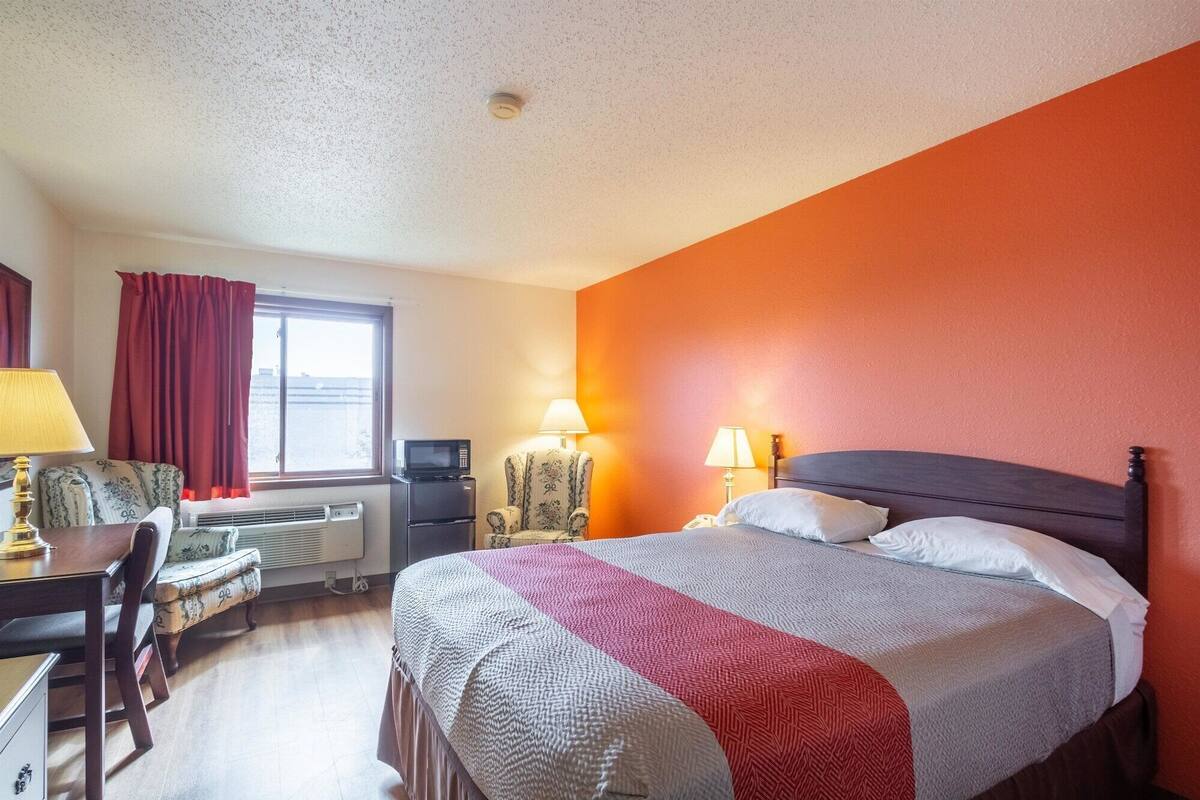 A hotel room with a bed, a desk, a chair, a window with curtains, and a radiator.