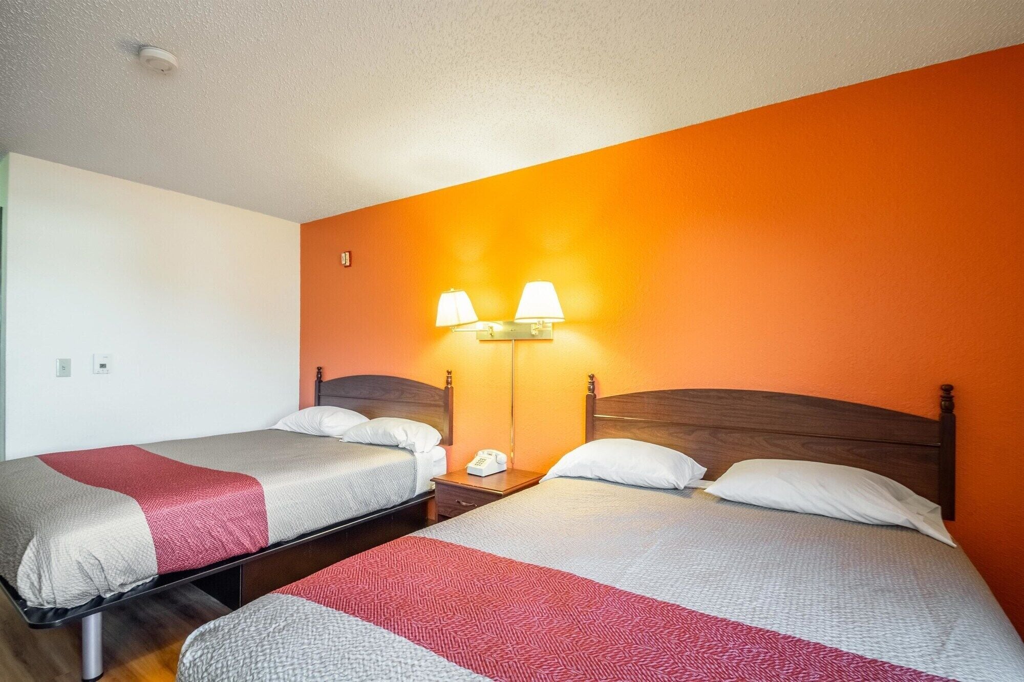 Une chambre d’hôtel avec deux lits, chacun doté d’une tête de lit en bois et d’une couvre-lit rouge et grise. Une table de chevet avec une lampe et un téléphone se trouve entre les deux lits.
