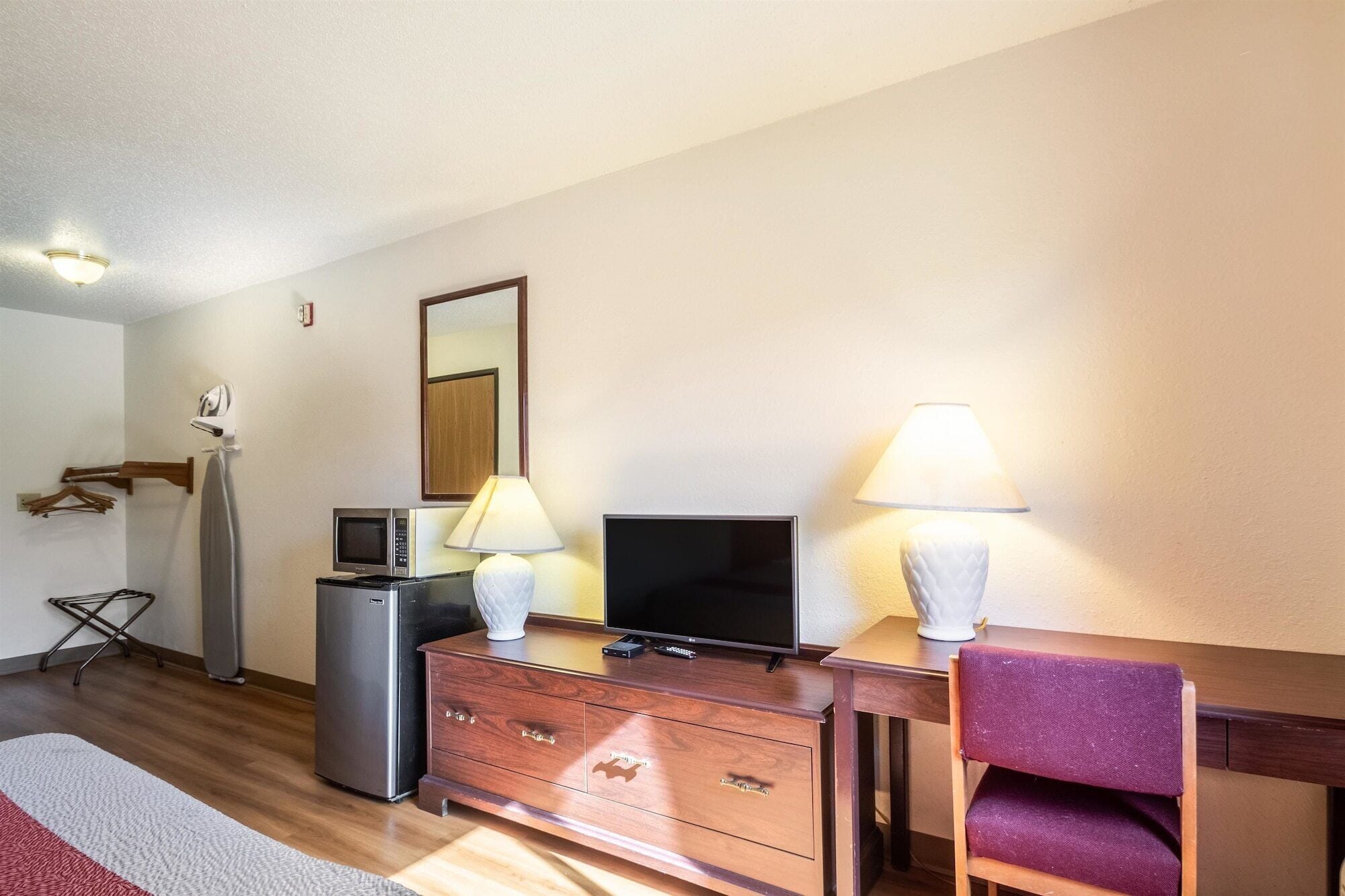 Een hotelkamer met een houten dressoir, een televisie, een magnetron, een koelkast, een bureau met stoel en een spiegel aan de muur.