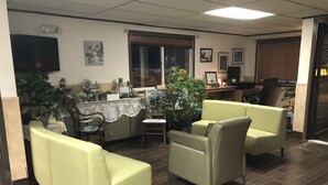 Un vestíbulo con mostrador de recepción, dos sillones verdes, una silla de madera, una planta en maceta y un televisor montado en la pared.