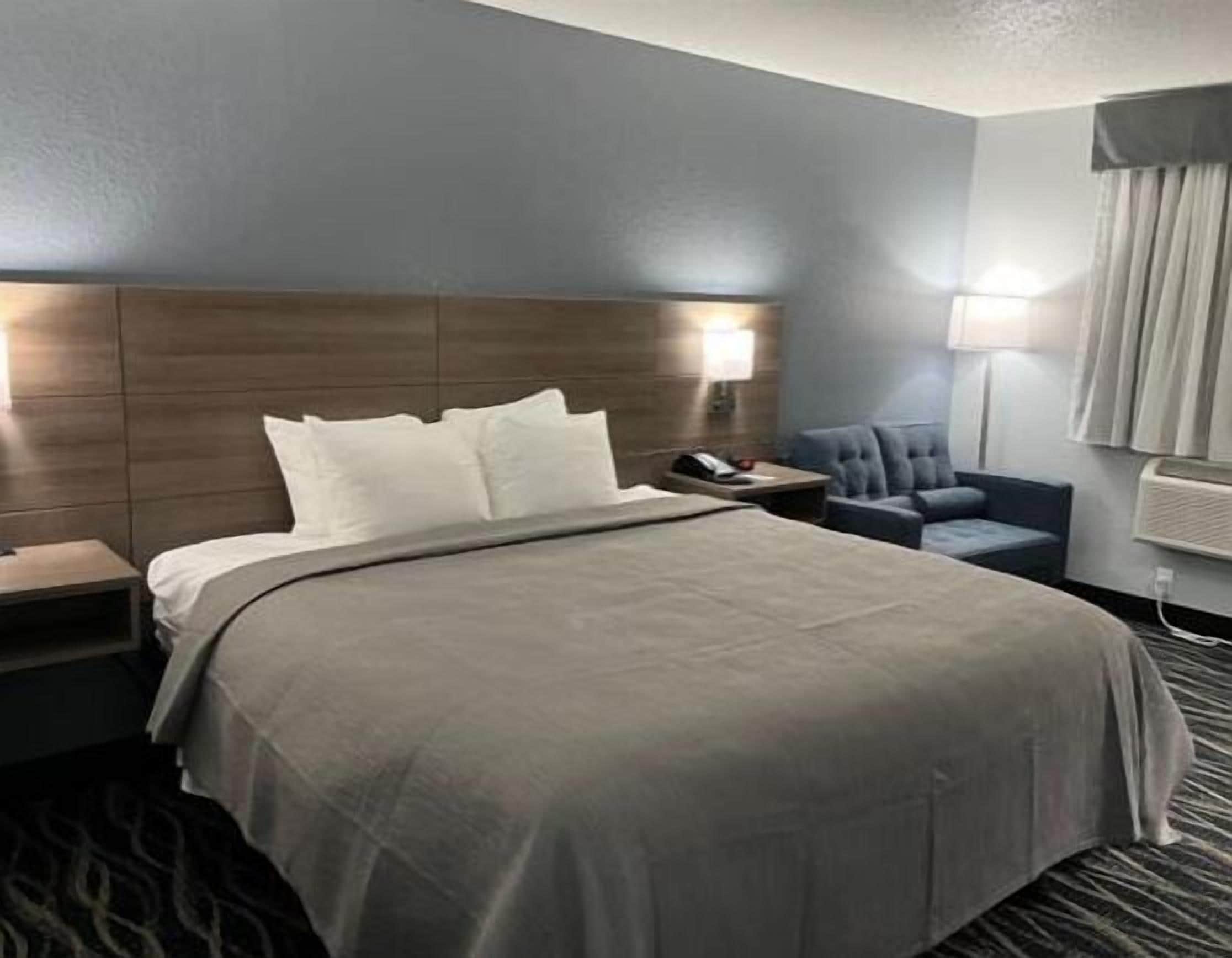 Hotellihuone, jossa on suuri sänky, yöpöytä lampun kanssa, tuoli ja ikkuna, jossa on verhot.