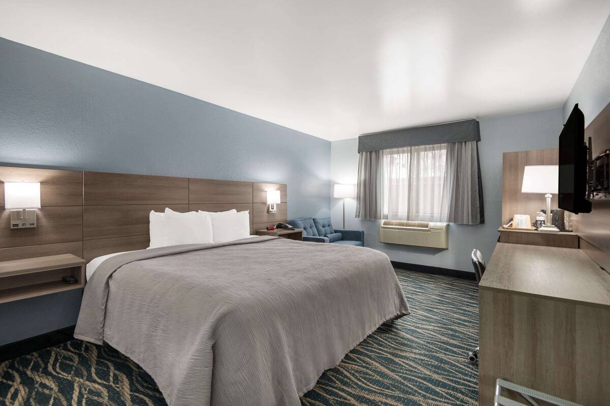 A hotel room with a large bed, bedside tables, a desk, a chair, a TV, and a window with curtains.