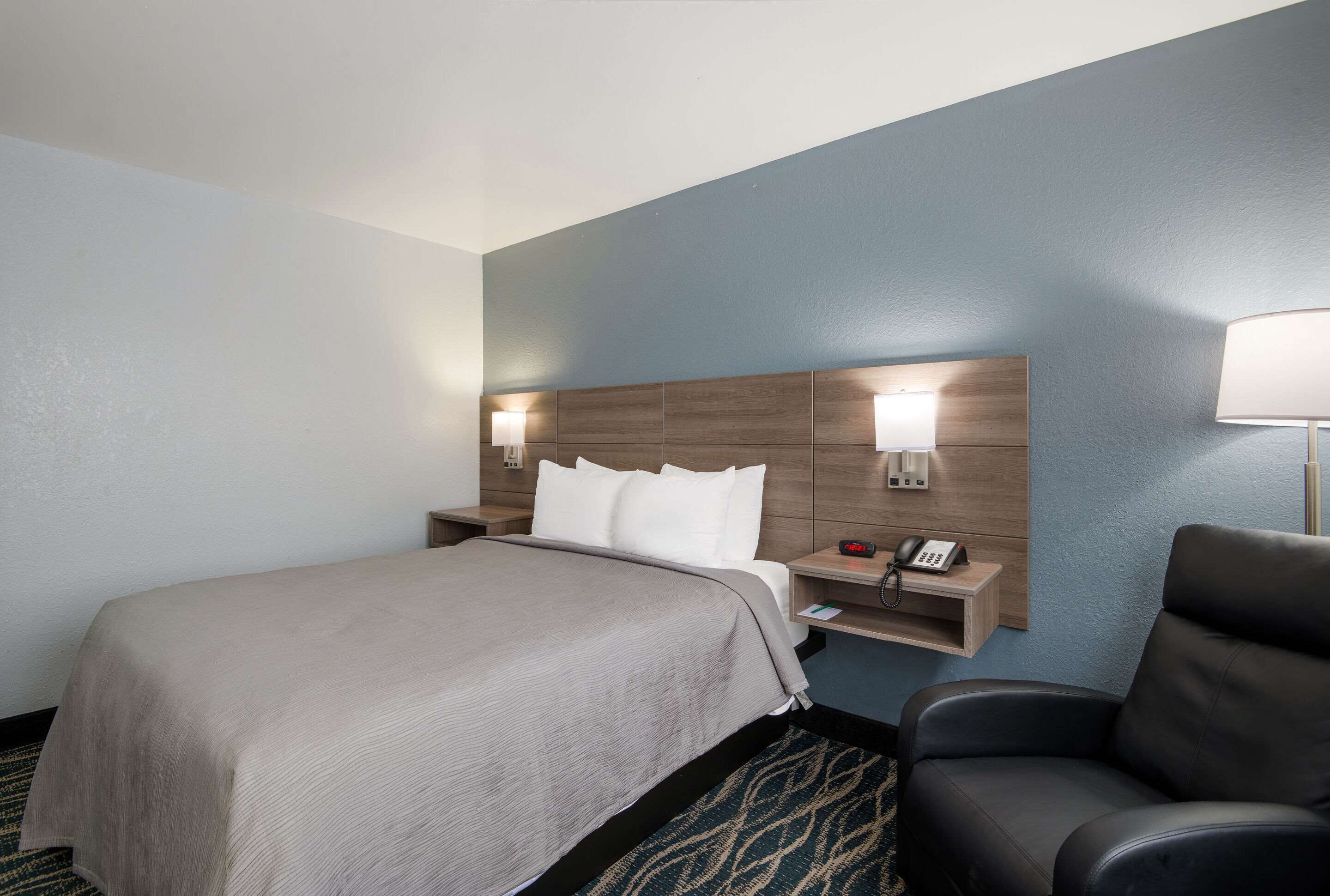 A hotel room with a large bed, a nightstand with a phone, a chair, and a wall-mounted lamp.