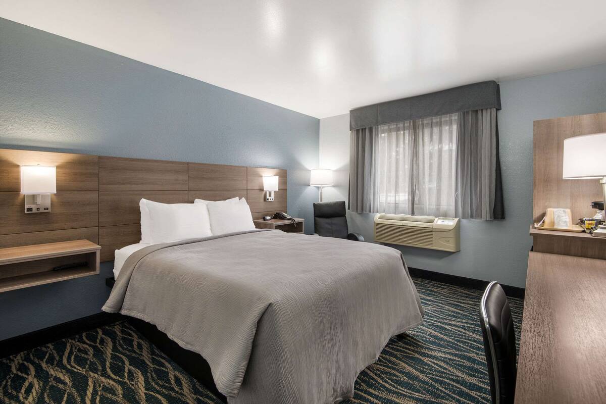 A hotel room with a large bed, bedside tables, a desk, a chair, a desk lamp, a window with curtains, and a wall-mounted air conditioning unit.
