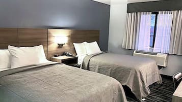 Quarto de hotel com duas camas, uma mesa de cabeceira com luminária, janela com cortinas e piso coberto por carpete.