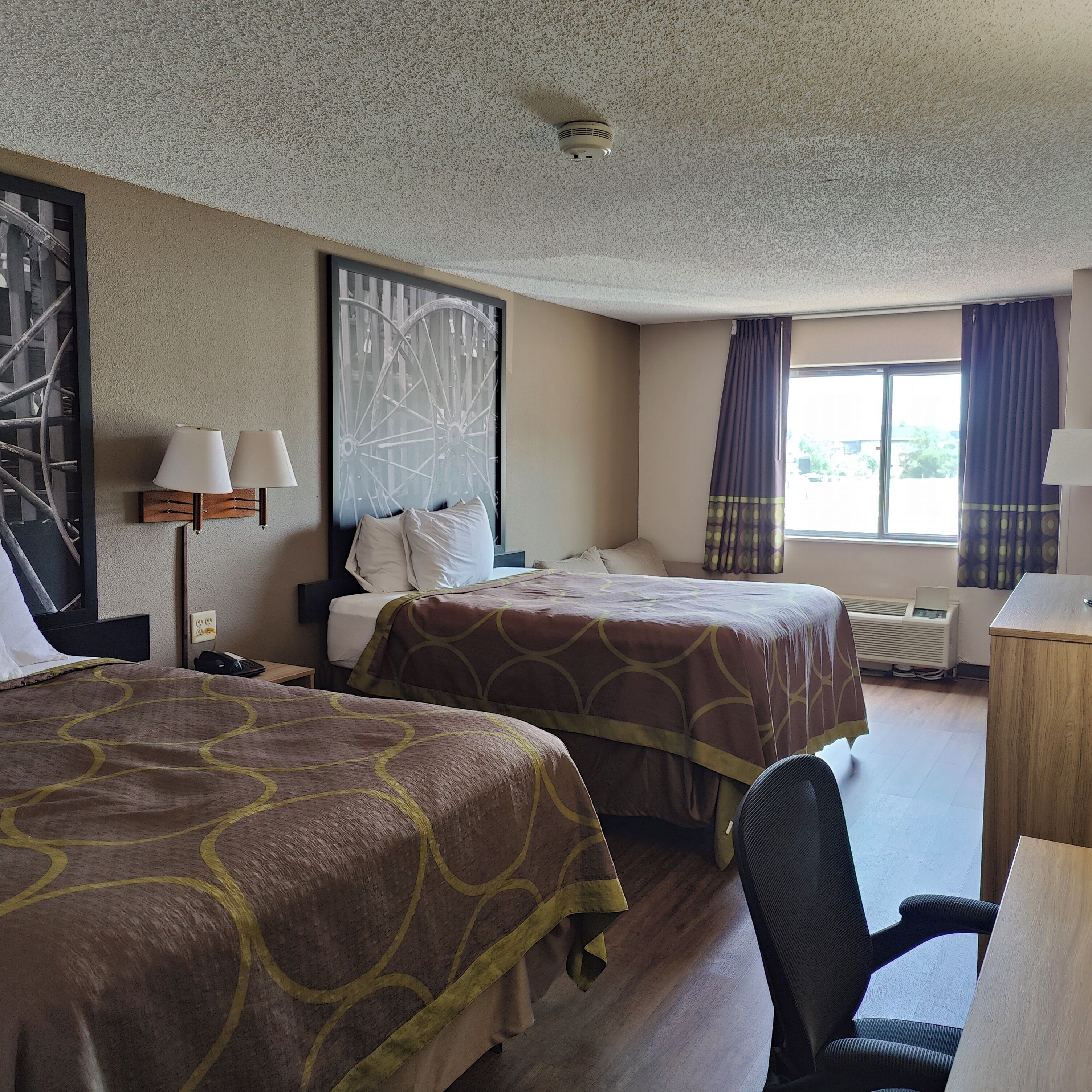 A hotel room with two beds, a desk, a chair, a radiator, and a window with curtains.
