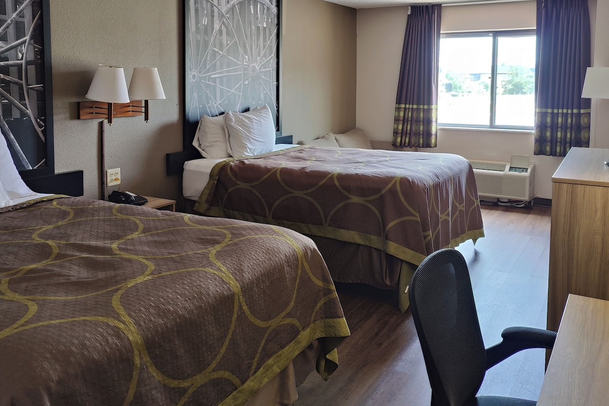 A hotel room with two beds, a desk, a chair, a radiator, and a window with curtains.