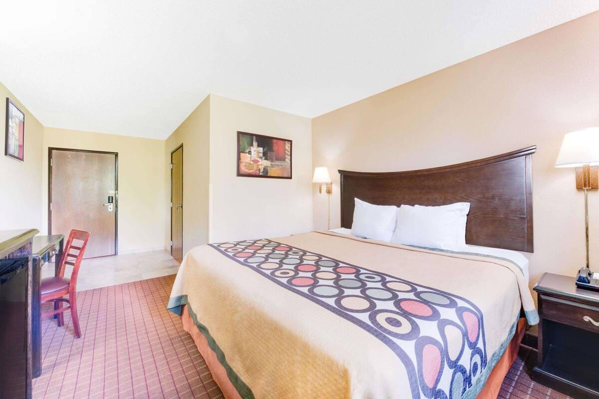 A hotel room with a bed, a desk, a chair, a door, and a wall-mounted lamp.