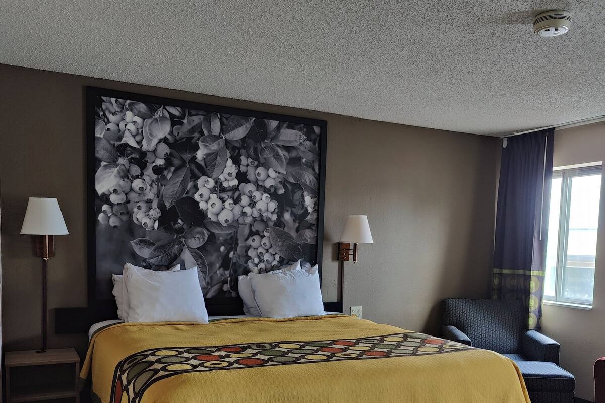 A hotel room with a large bed, two bedside lamps, a dark armchair, and a large floral artwork on the wall.