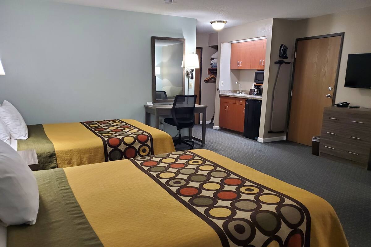 A hotel room with two beds, a desk, a chair, a kitchenette, and a TV.