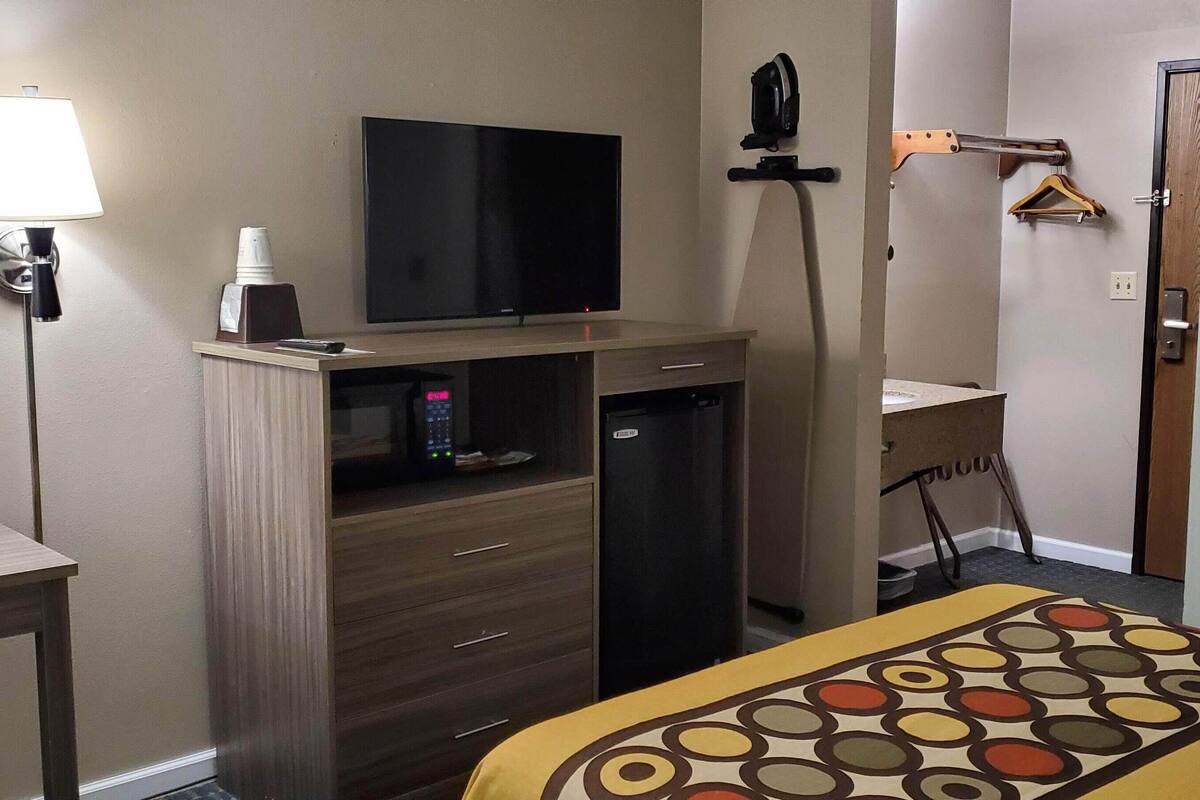 A hotel room with a bed, a desk with a lamp, a TV, a mini-fridge, and a coat rack.
