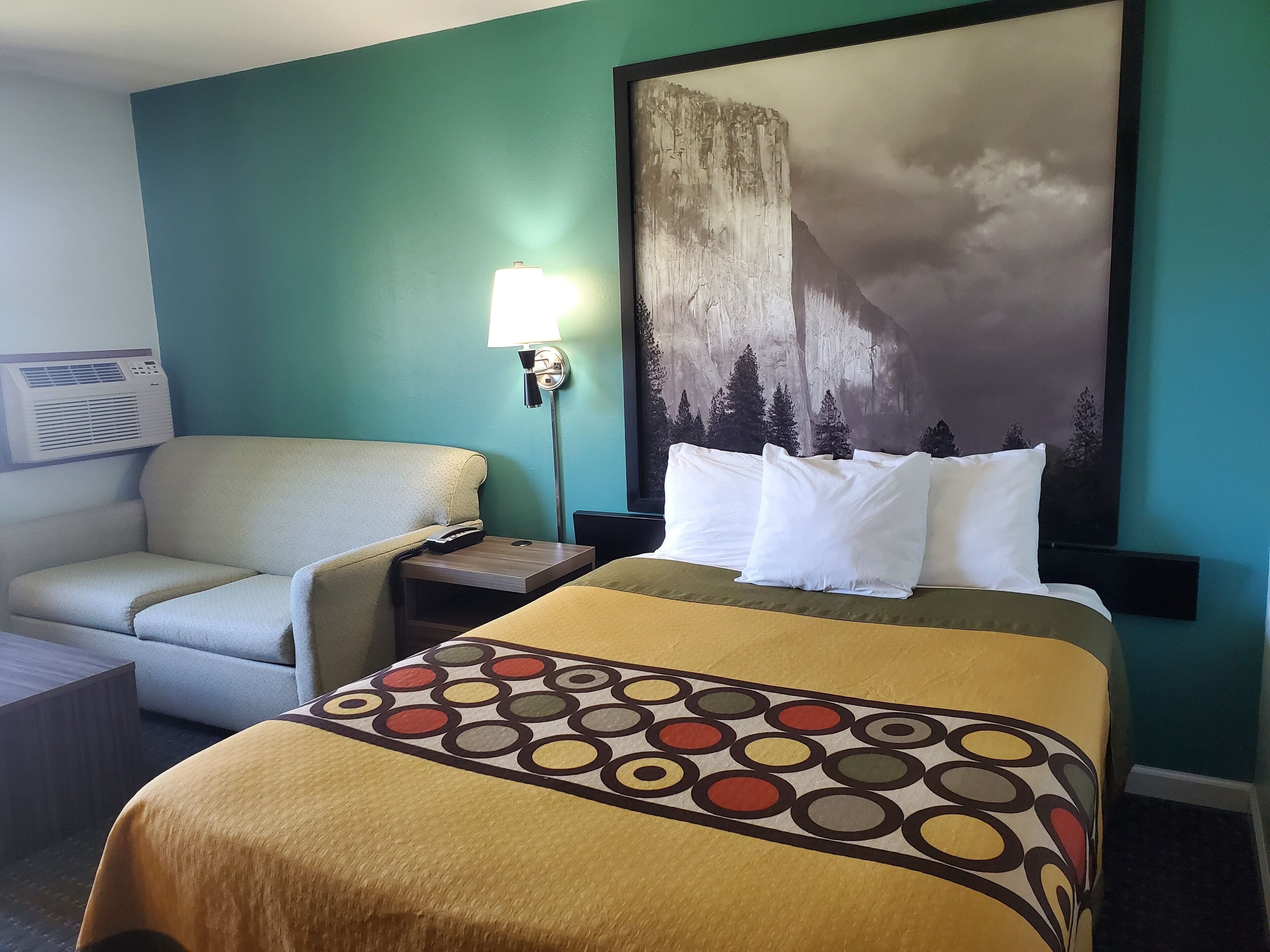 A hotel room with a bed, a chair, a nightstand, a lamp, and a painting of a mountain.