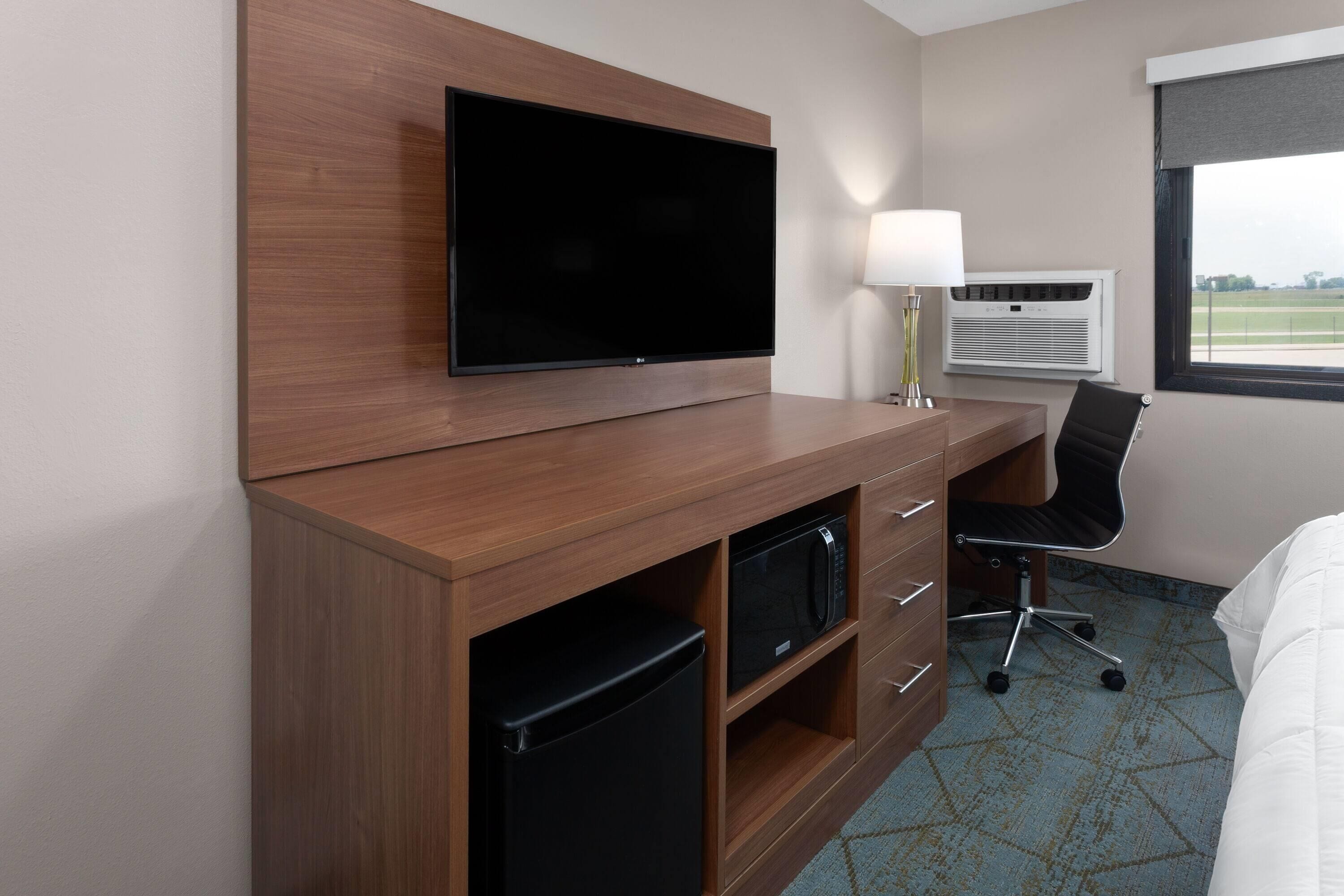 A hotel room with a flat-screen TV mounted on a wooden cabinet, a desk with a chair, a lamp, an air conditioning unit, and a bed with a white duvet.