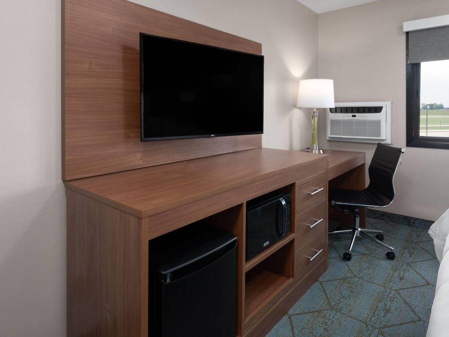 A hotel room with a flat-screen TV mounted on a wooden cabinet, a desk with a chair, a lamp, an air conditioning unit, and a bed with a white duvet.