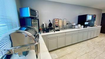 Cocina de hotel con estación de desayuno autoservicio, que incluye una máquina para gofres, una cafetera y varios dispensadores de alimentos.