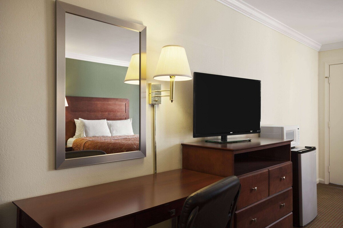 A hotel room with a wooden desk, a flat-screen TV, a lamp, and a mirror reflecting a bed with white bedding.