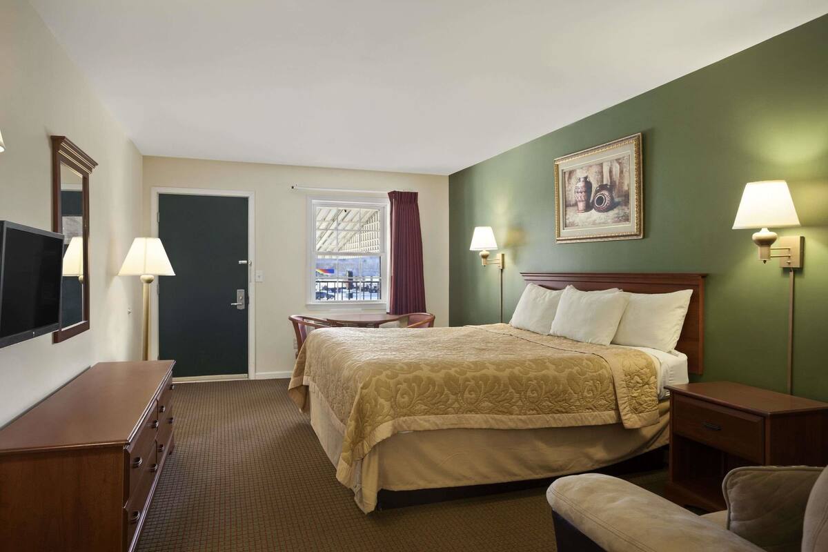 Habitación de hotel con cama, lámparas de noche, televisor, cómoda y ventana con cortinas.