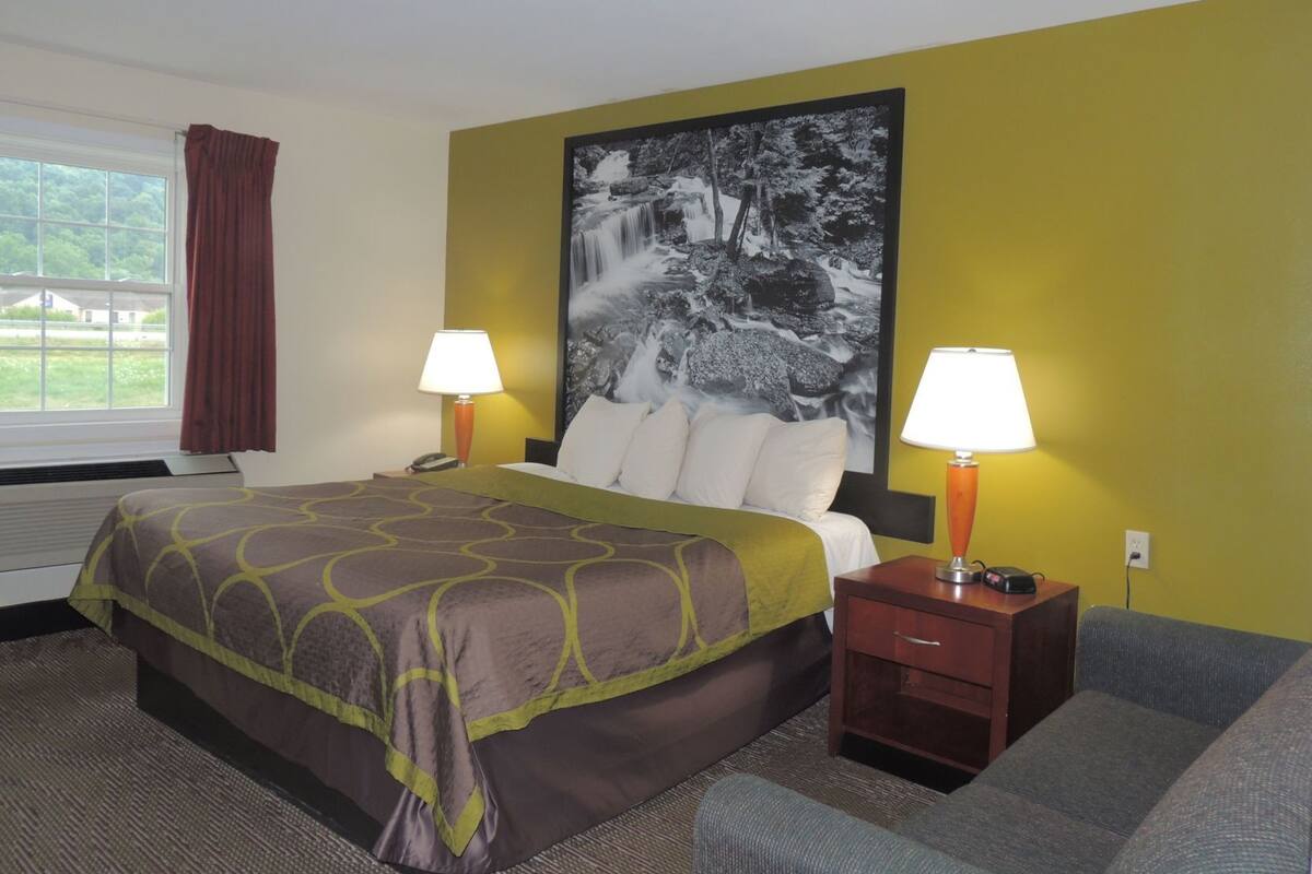 Una habitación de hotel con una cama, dos lámparas de noche, una mesita de noche, un sofá, un cuadro grande en la pared y una ventana con cortinas.