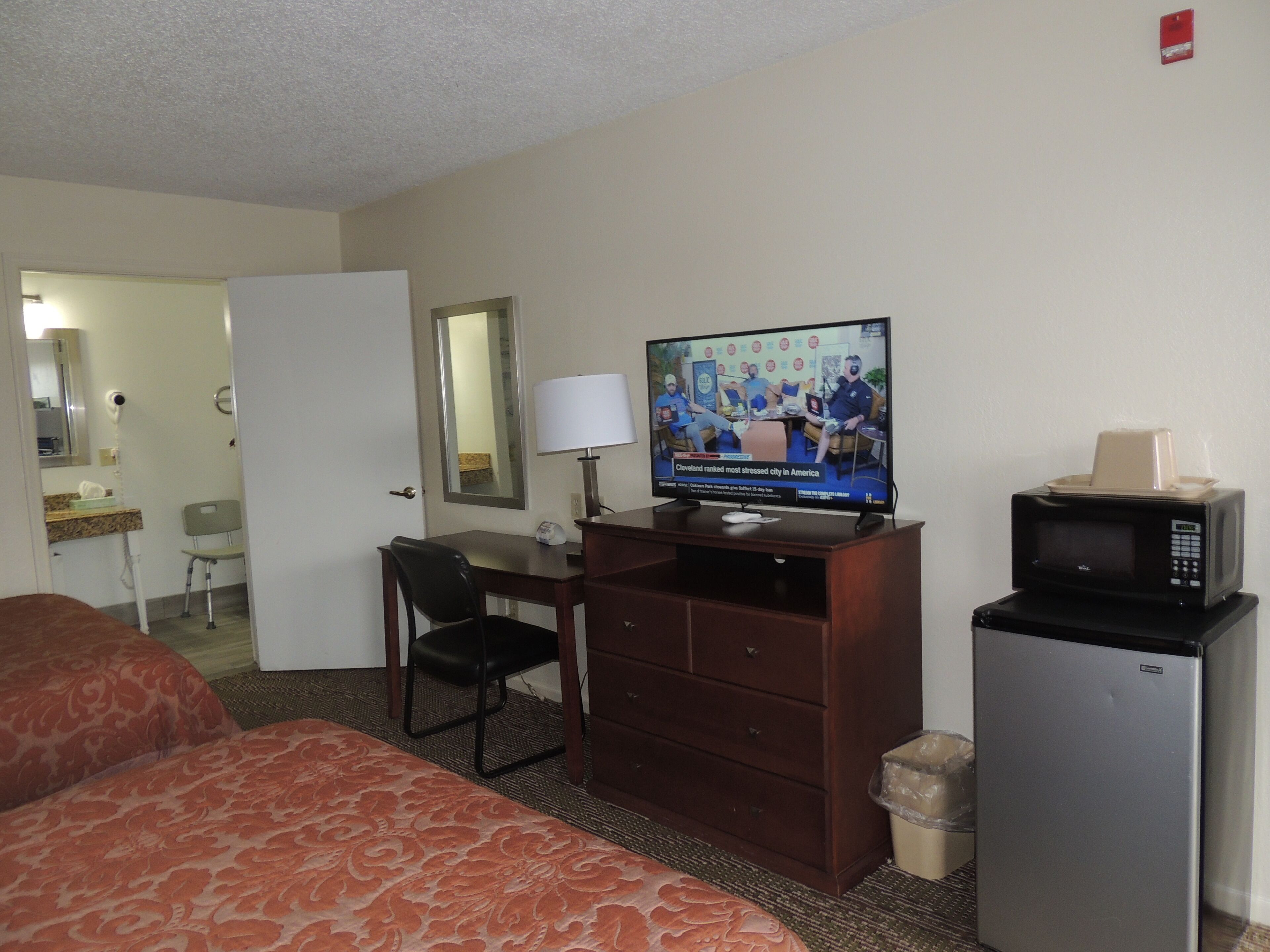 A hotel room with a bed, a desk with a chair, a television, a microwave, and a trash can.